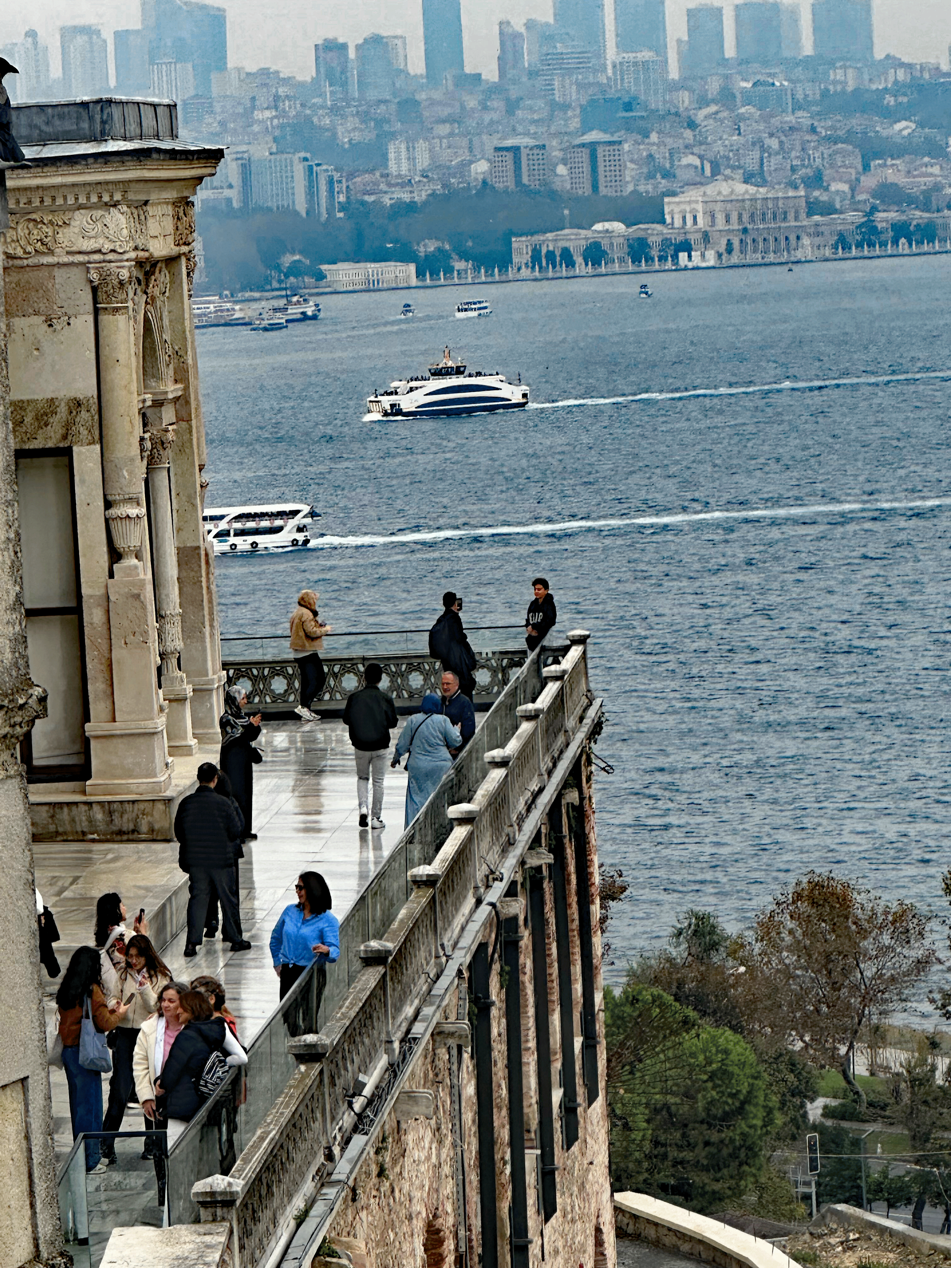 View of Istanbul's Bosphorus Strait with people on a terrace, modern skyline in the background, and boats on the water.