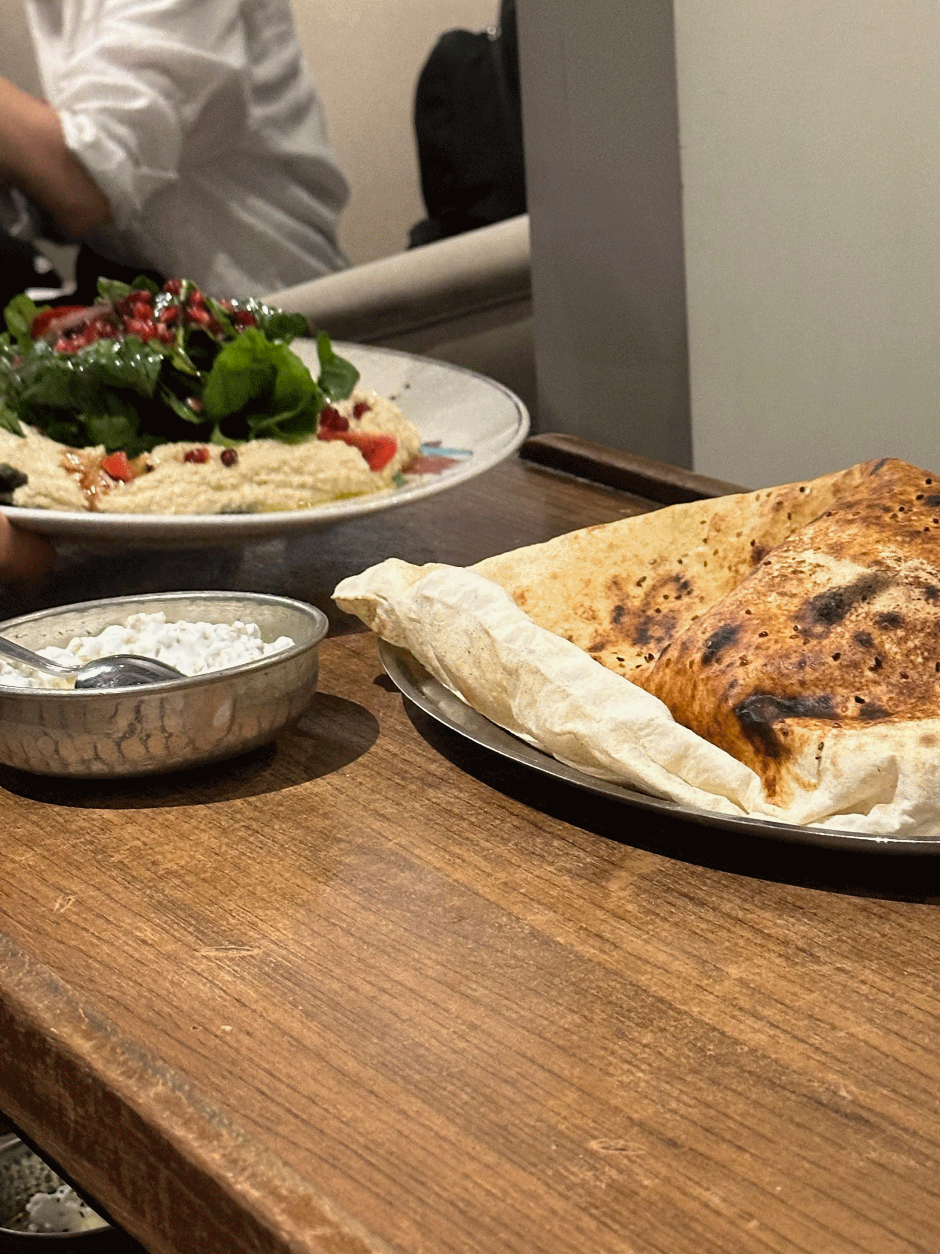 A plate of Turkish salad with pomegranate seeds and spinach next to a serving of flatbread and a bowl of yogurt on a wooden table.