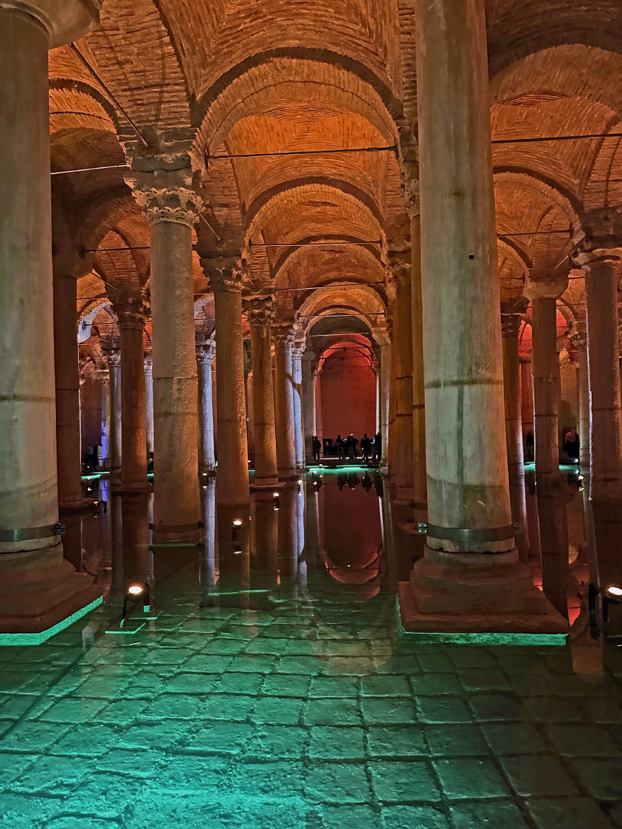 Interior view of the Basilica Cistern in Istanbul, featuring a series of arched stone columns reflected in water with colorful lighting.