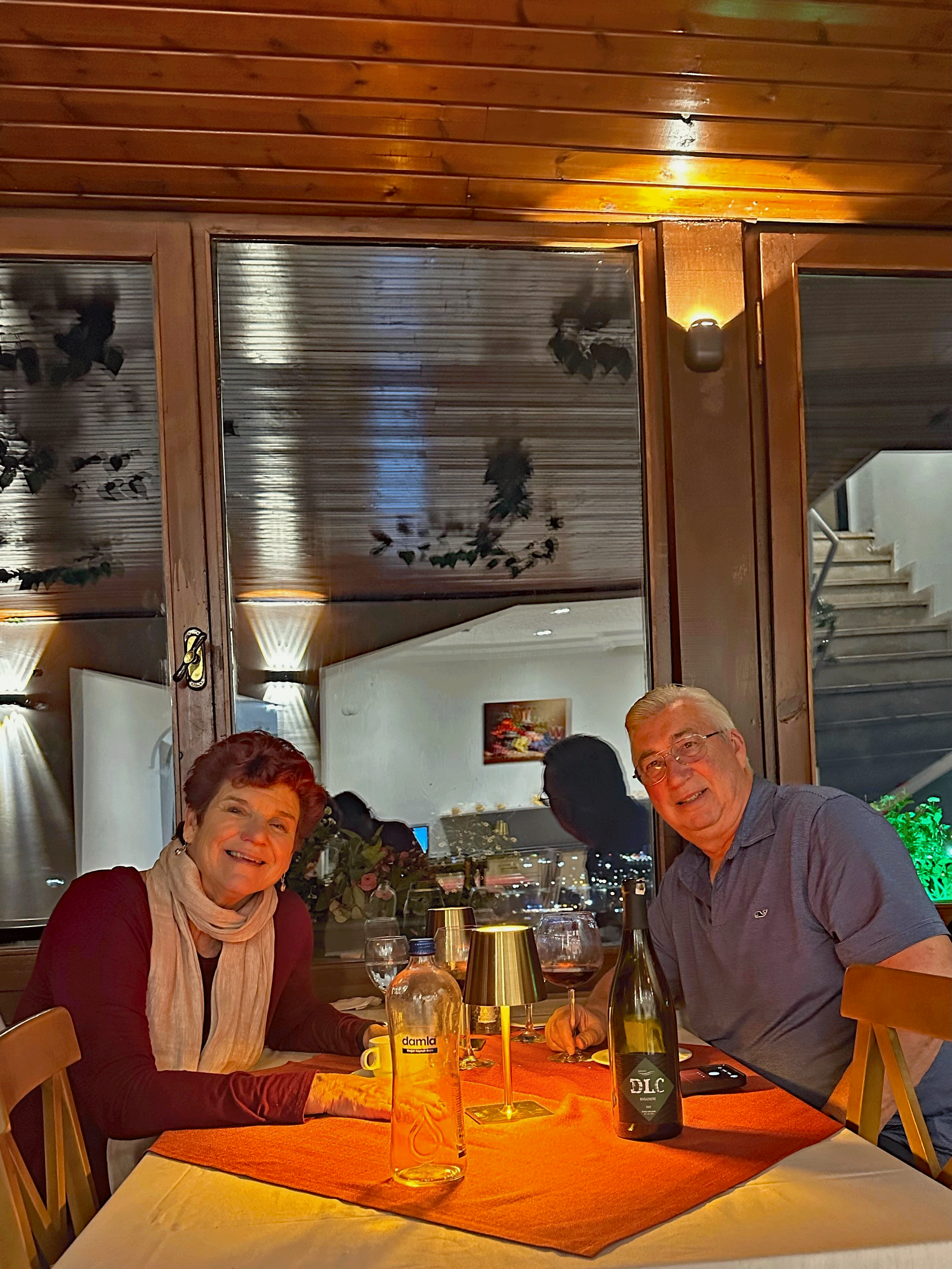 A couple dining at a cozy table in a warmly lit restaurant, with wine glasses and a bottle visible on the table.