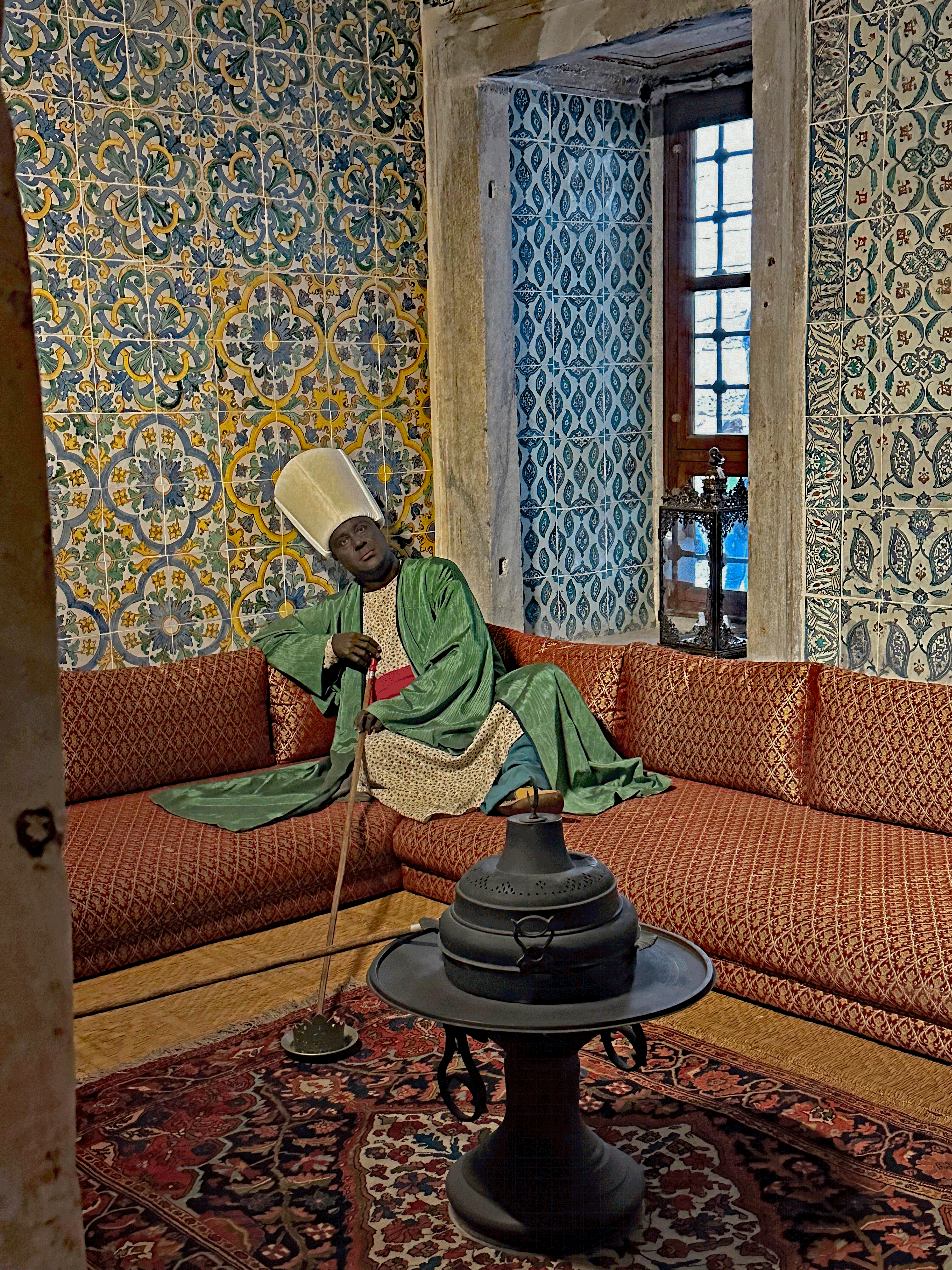 A historical recreation of a figure in traditional Ottoman clothing seated in a room adorned with patterned tiles and a intricately designed couch.