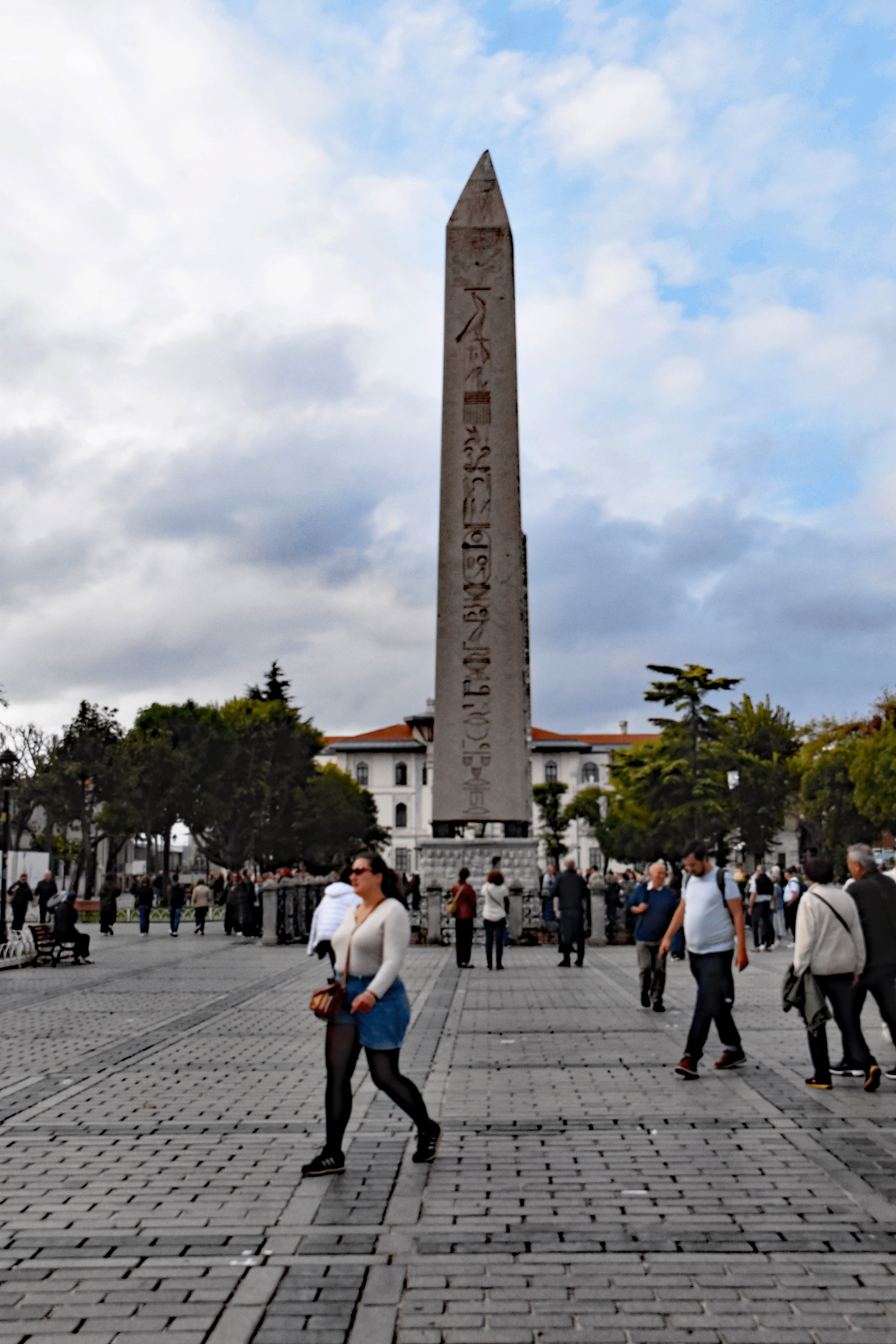 A busy square in Istanbul featuring a tall ancient obelisk surrounded by pedestrians strolling on a paved walkway under a cloudy sky.