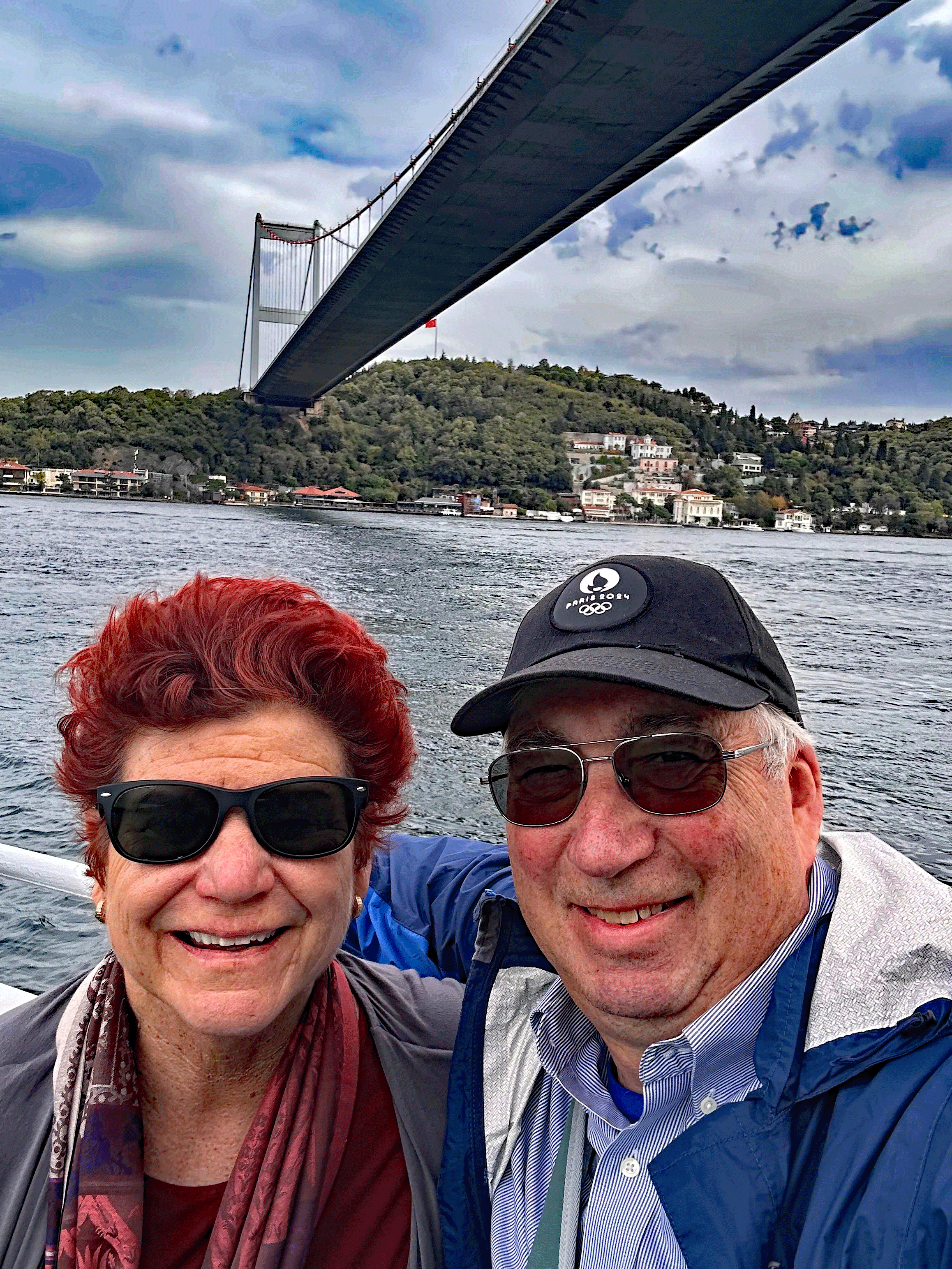 A couple smiling and posing for a selfie on a boat with a large bridge in the background. The scene depicts a waterway surrounded by greenery and buildings along the shore, under a partly cloudy sky.