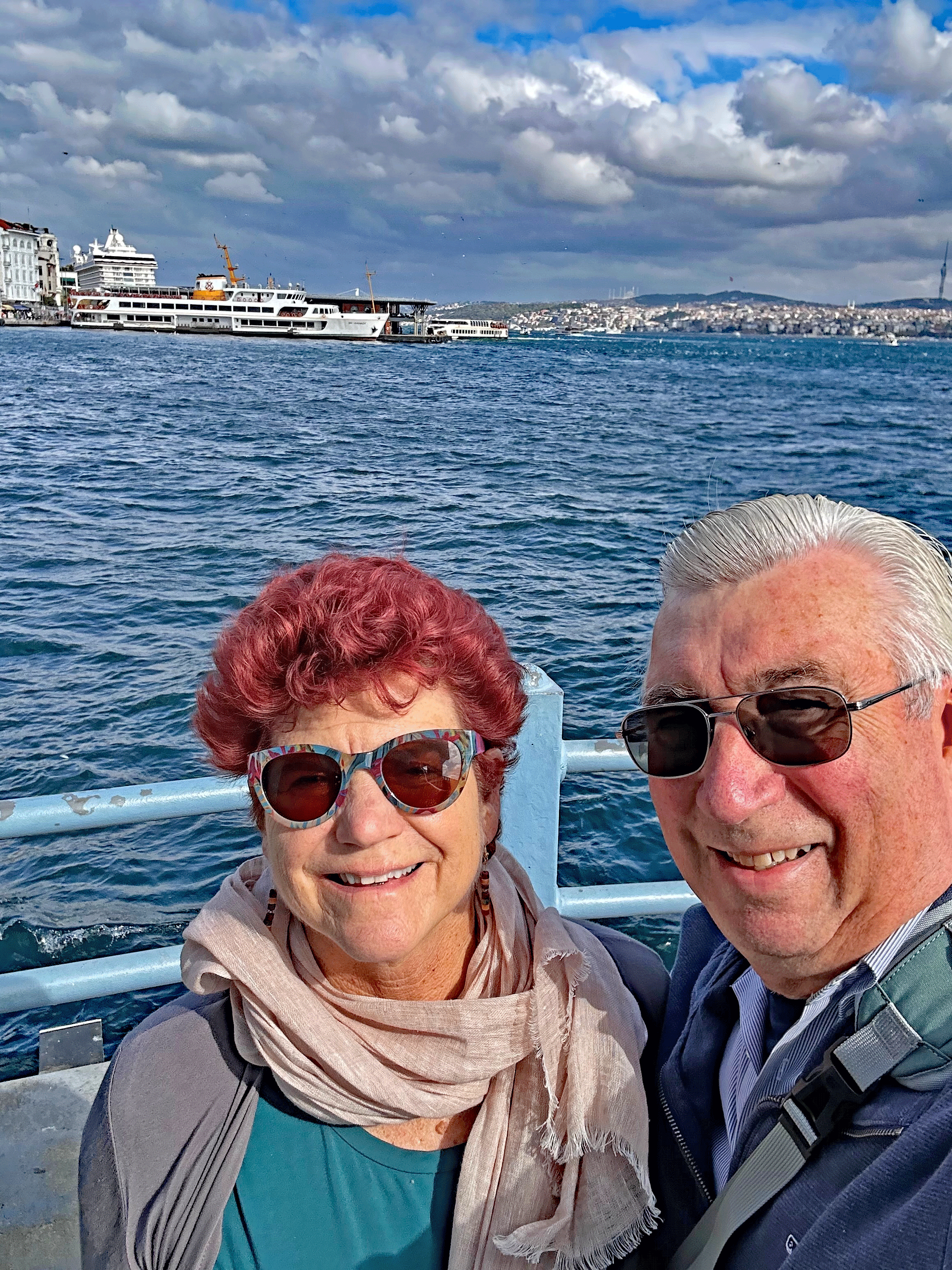 A couple posing for a selfie at the waterfront in Istanbul, with boats and a cloudy sky in the background.