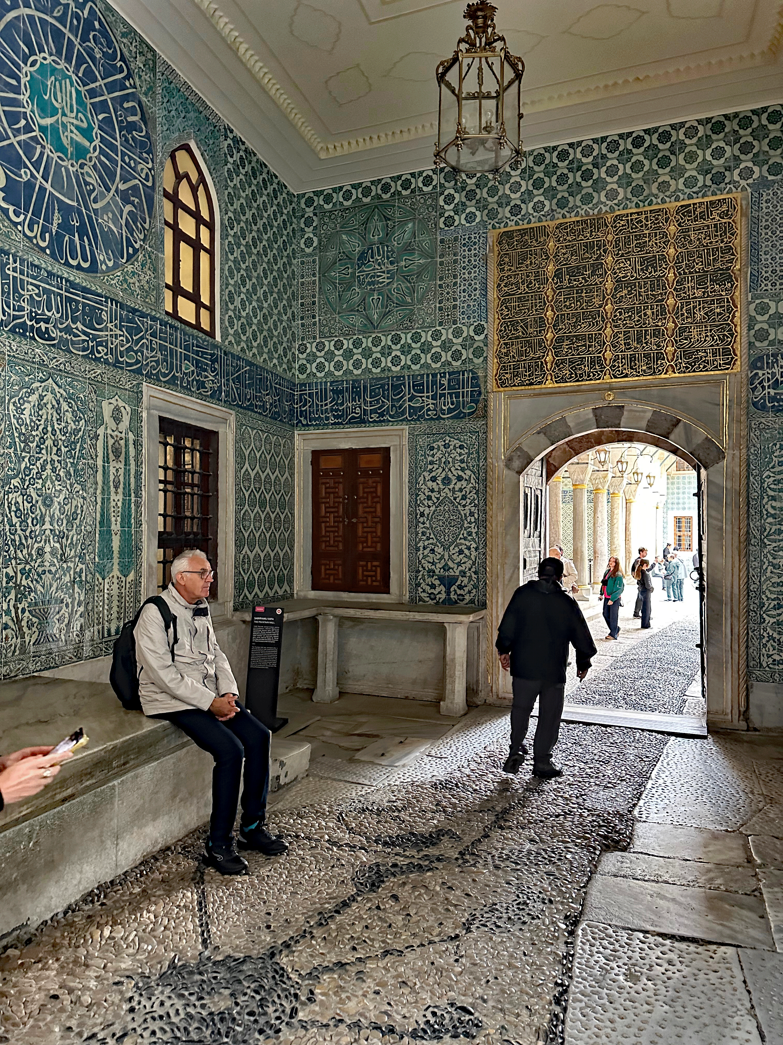 Interior view of a room in Topkapi Palace featuring intricate blue tile work and calligraphy on the walls, with visitors exploring and one person seated on a stone bench.