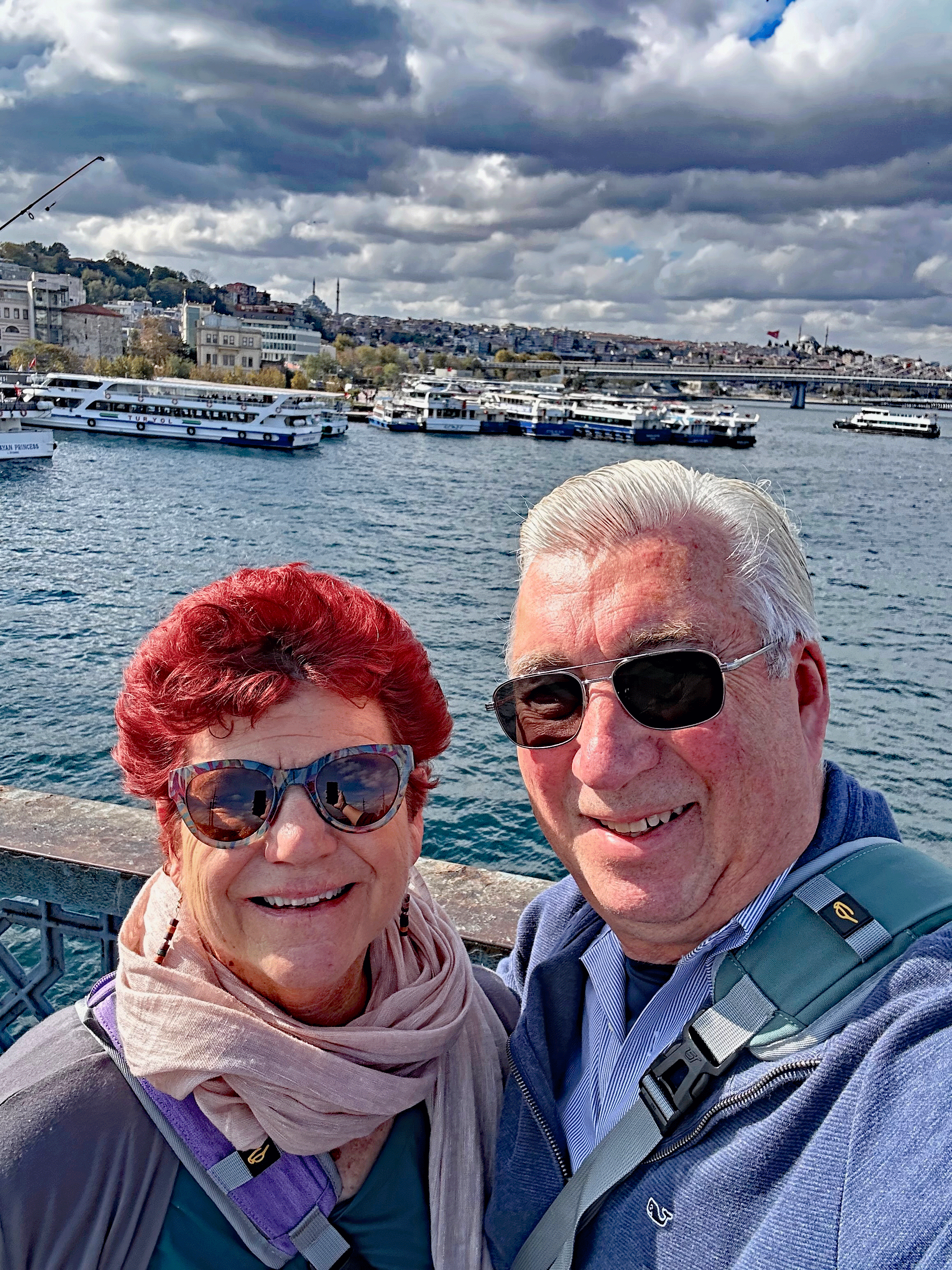 A couple posing for a selfie on a bridge in Istanbul, with the skyline and boats in the background under a cloudy sky.