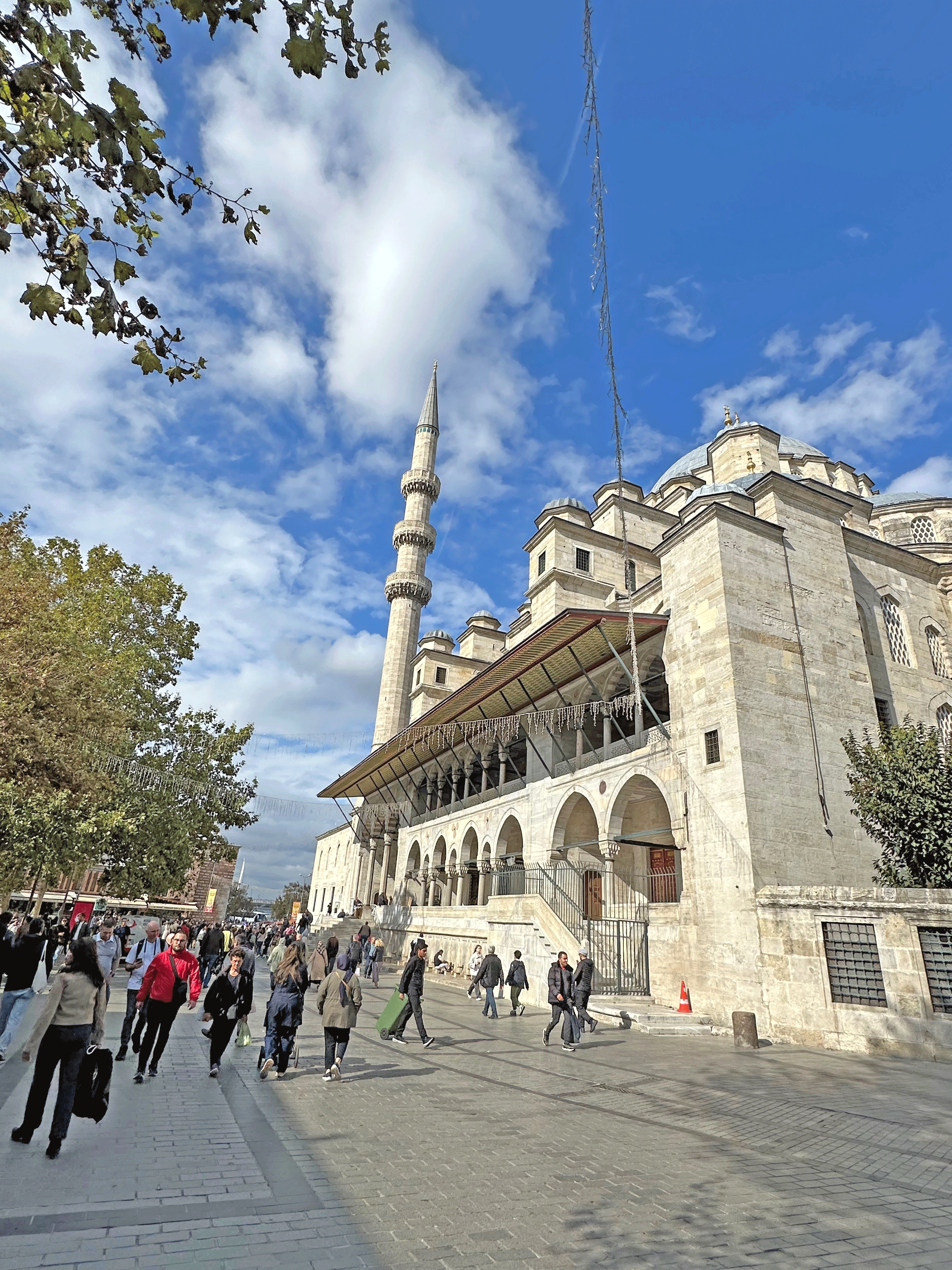 A bustling street scene in Istanbul with a prominent mosque in the background, showcasing its architectural details under a blue sky with clouds.