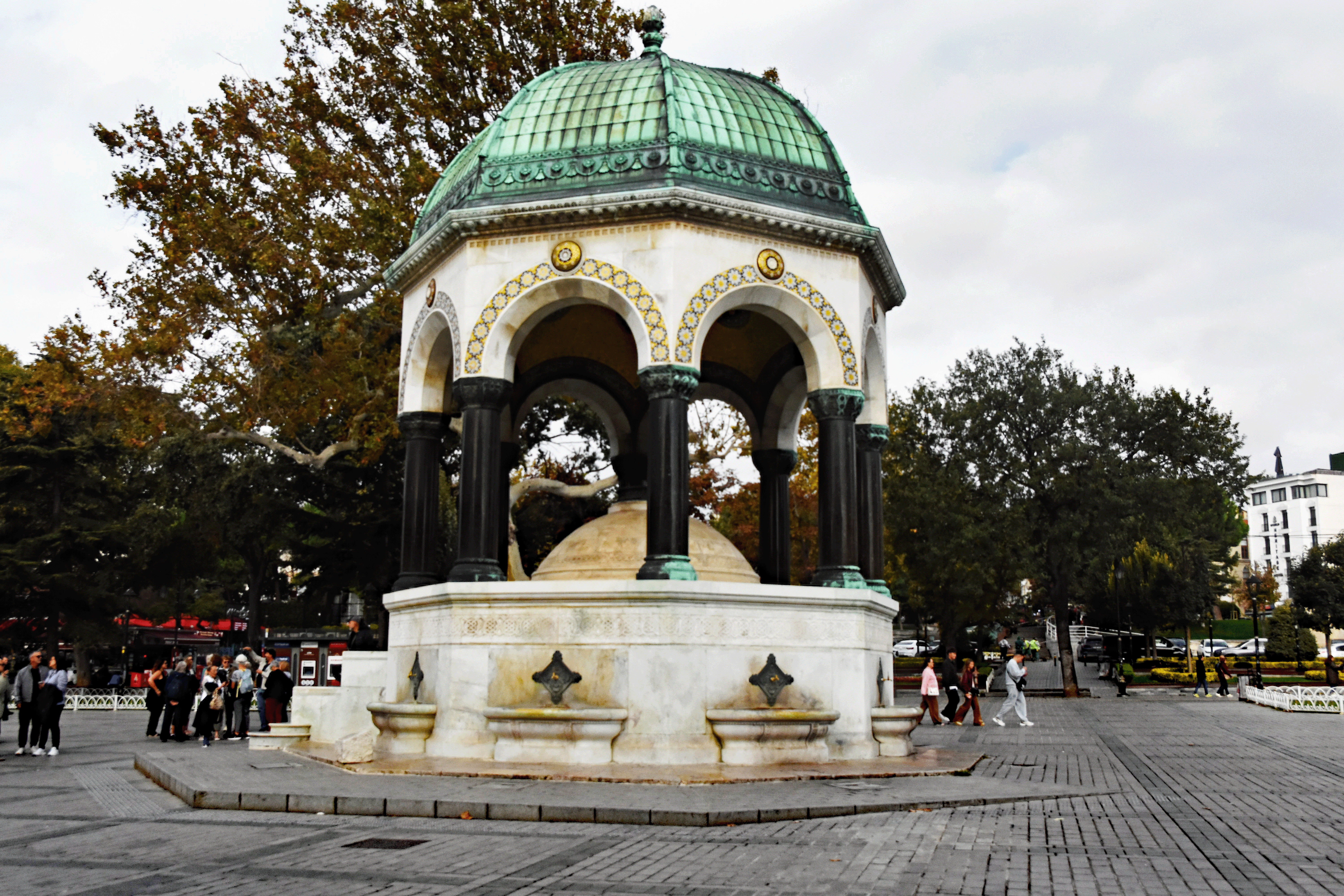 A historical fountain with a green domed roof and ornate architectural details, surrounded by trees and people in a public square in Istanbul.