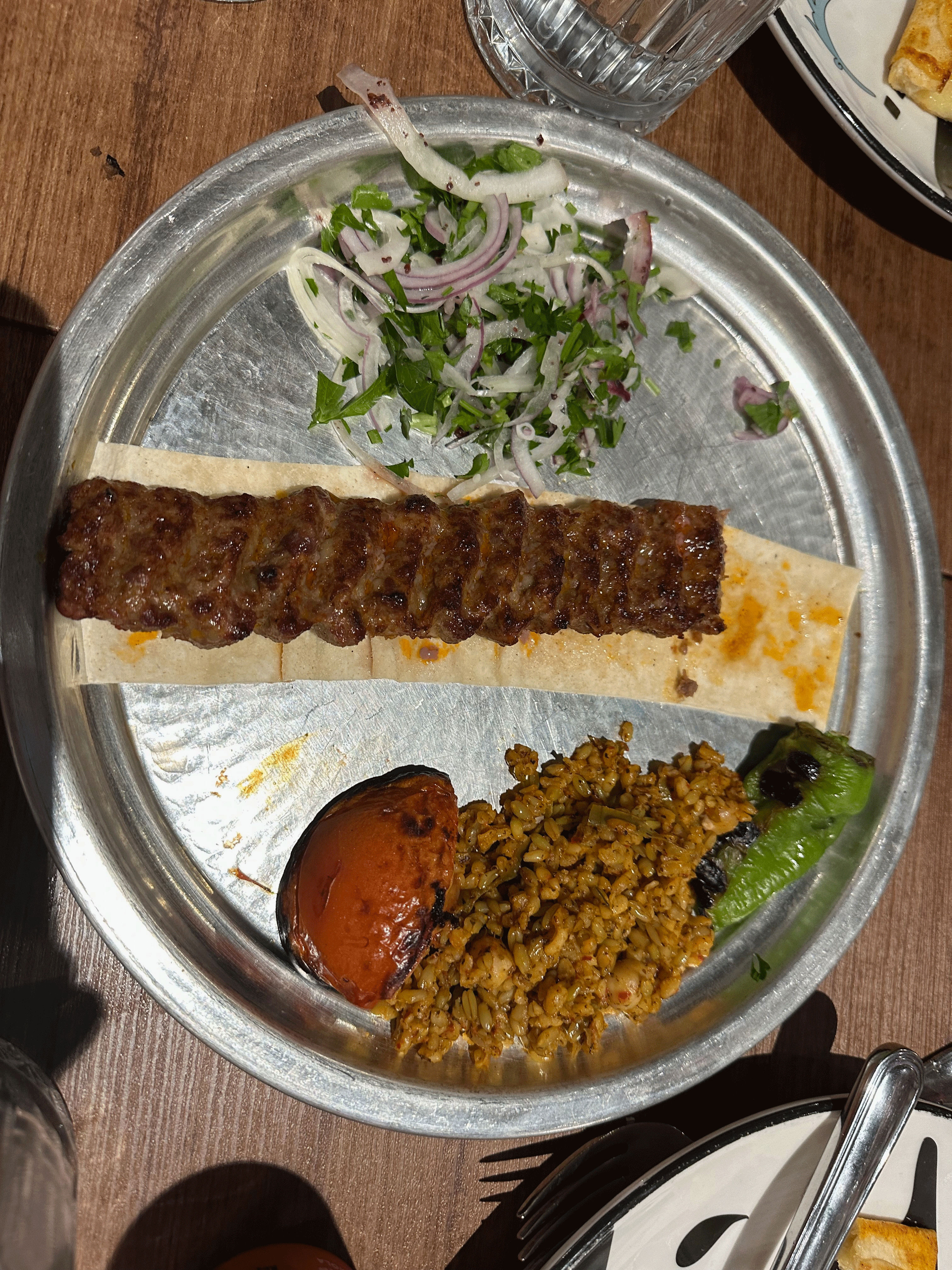 A plate of Turkish kebab served on flatbread with grilled vegetables, rice, and fresh herbs.