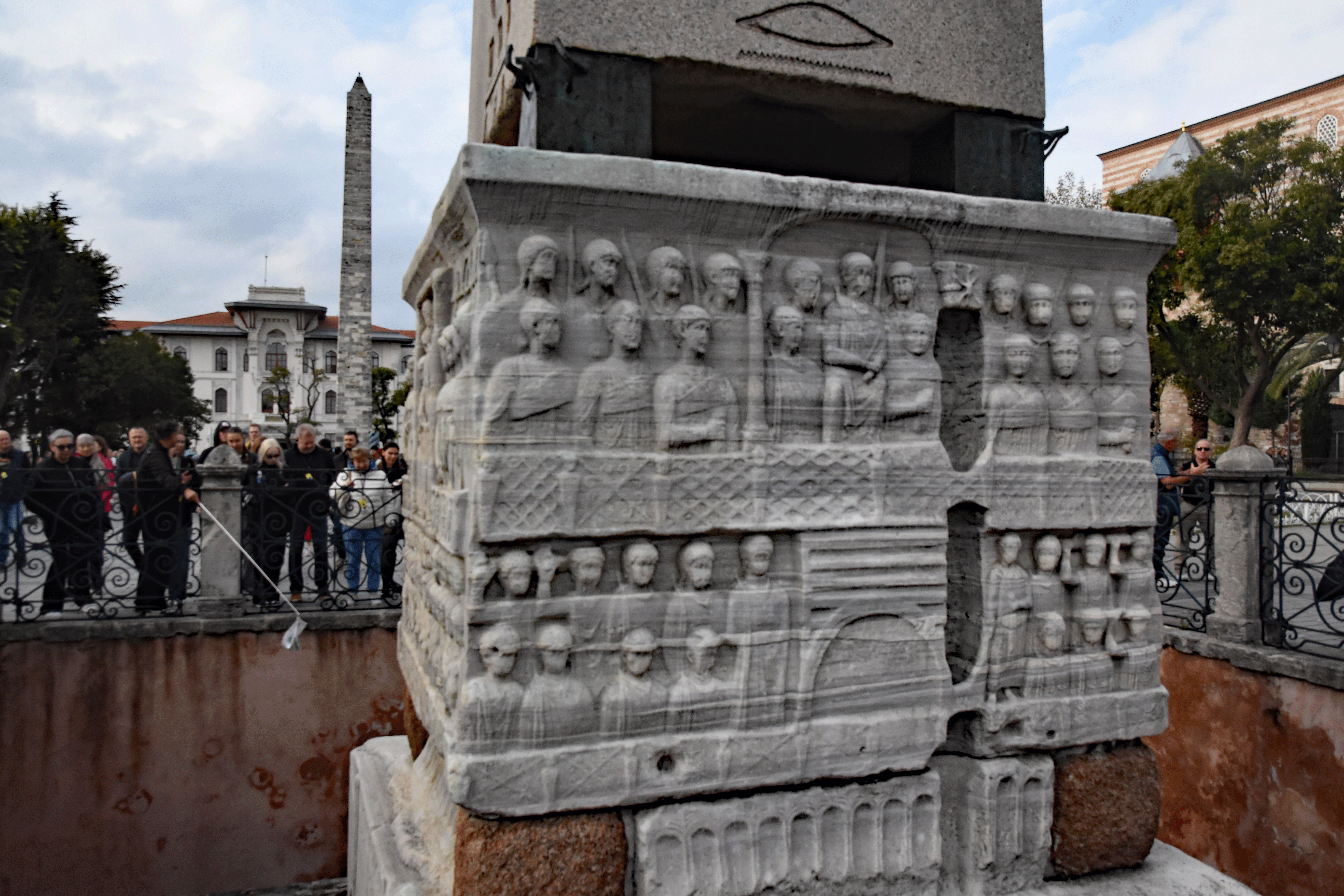 An ancient stone monument with intricate carvings and reliefs, surrounded by a crowd of tourists, featuring the obelisk of Theodosius in the background.