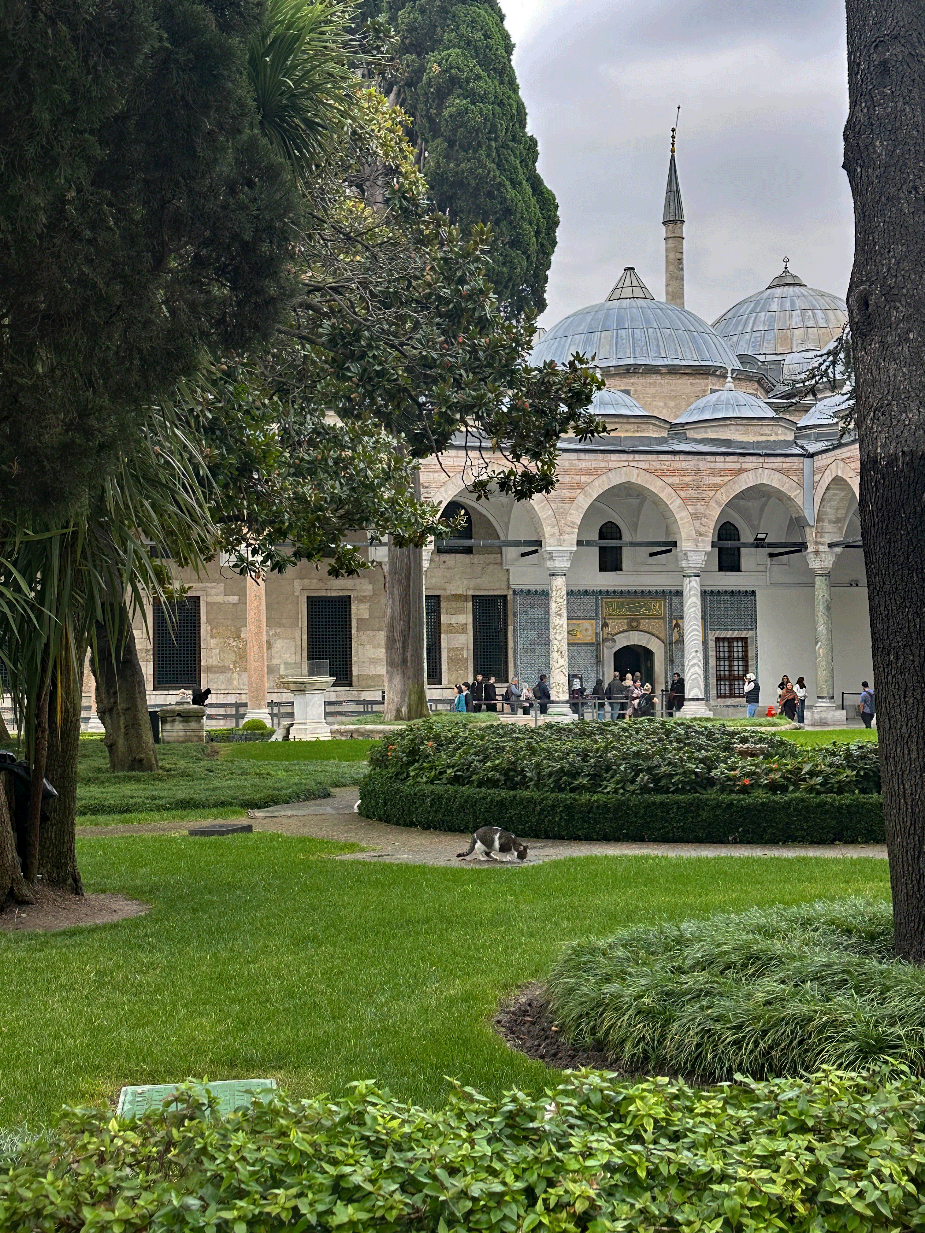 A view of the Topkapi Palace gardens featuring lush greenery, neatly trimmed bushes, and visitors walking in the background, with ornate domes and a minaret visible beyond the trees.