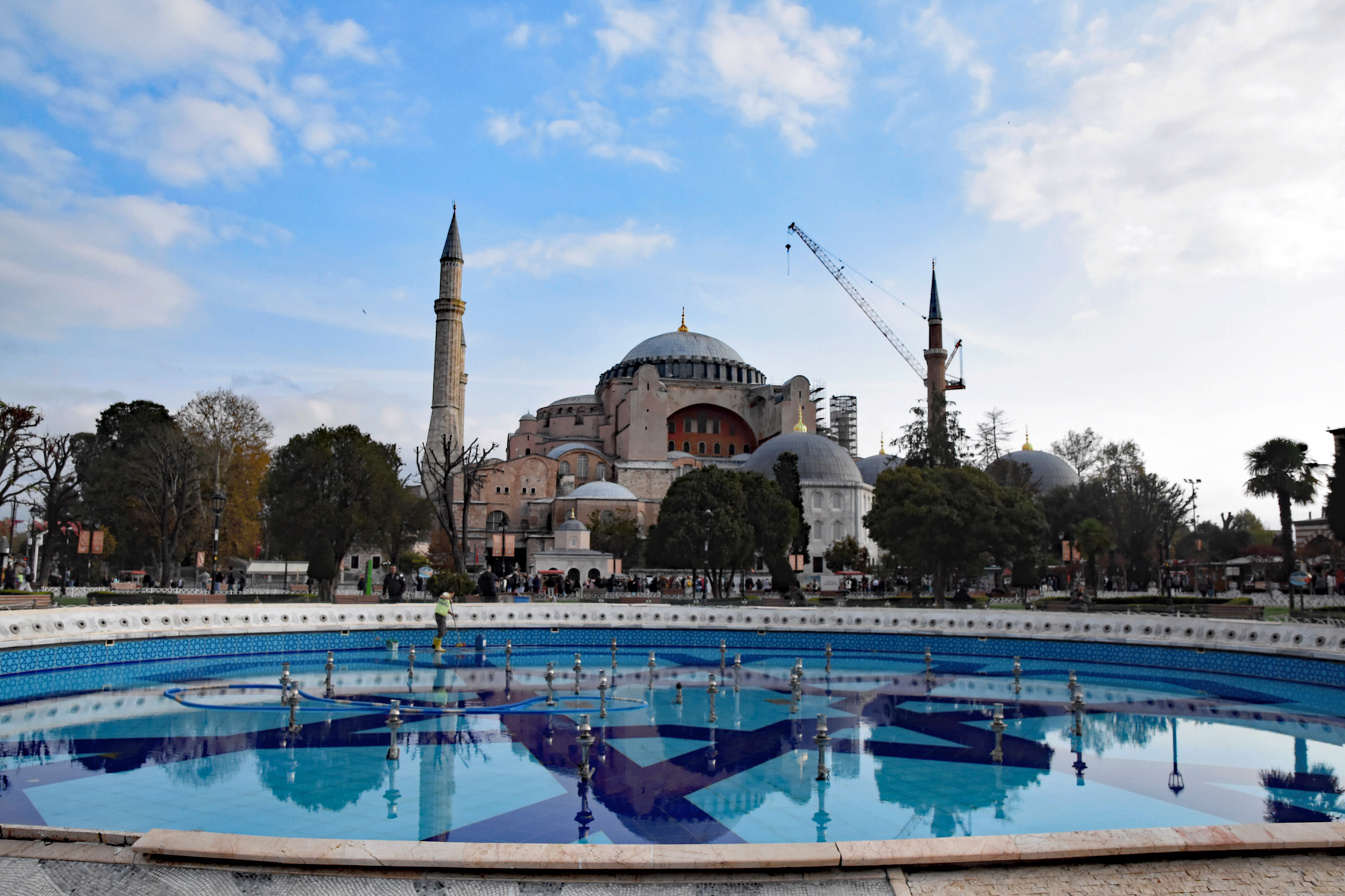 View of the Hagia Sophia against a cloudy sky, featuring a fountain in the foreground and construction cranes beside the mosque.