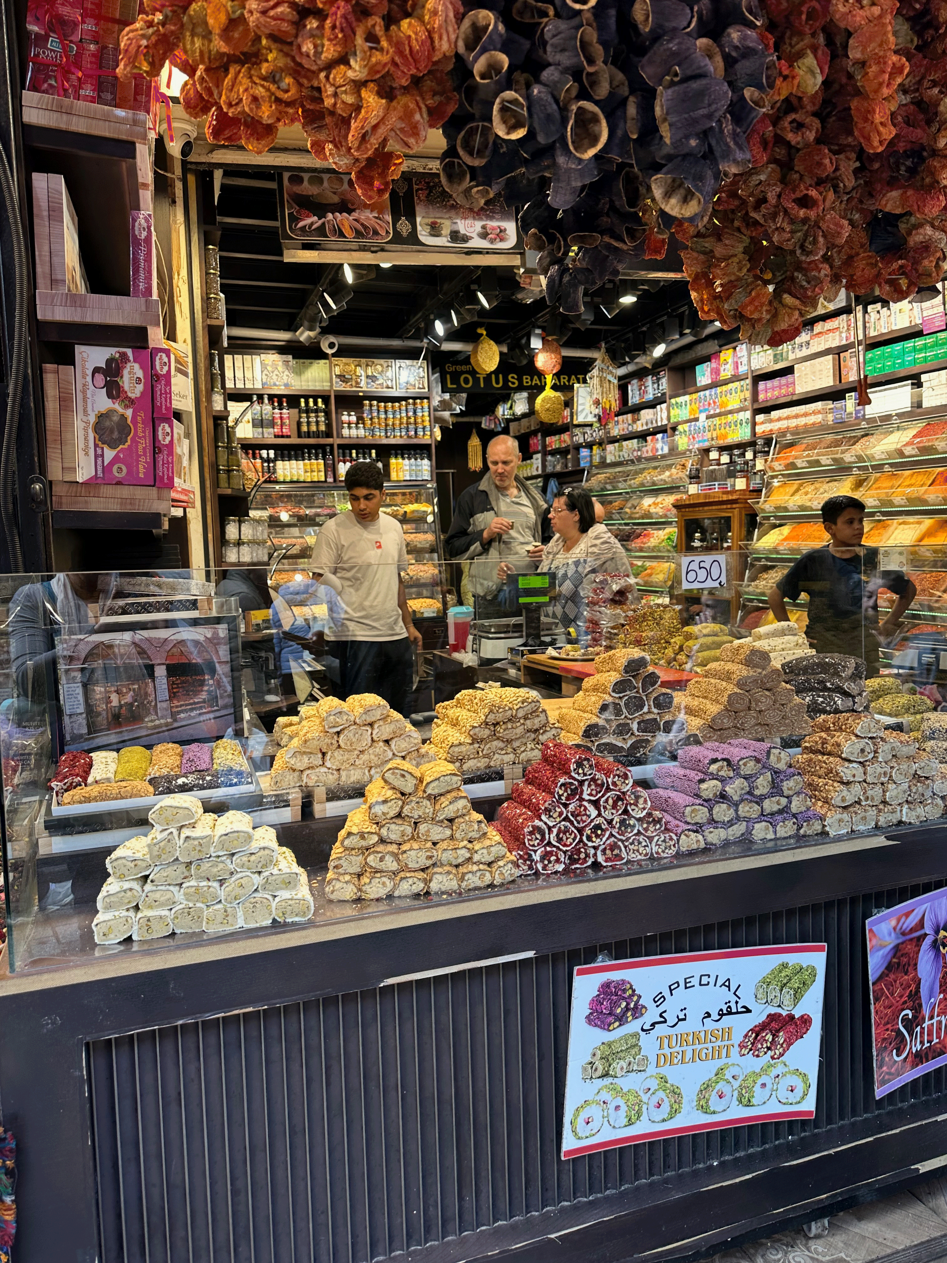A vibrant shop in the Spice Market in Istanbul, displaying various types of Turkish delight arranged in decorative pyramids. Dried fruits hang from the ceiling, and shoppers engage with the shopkeepers.