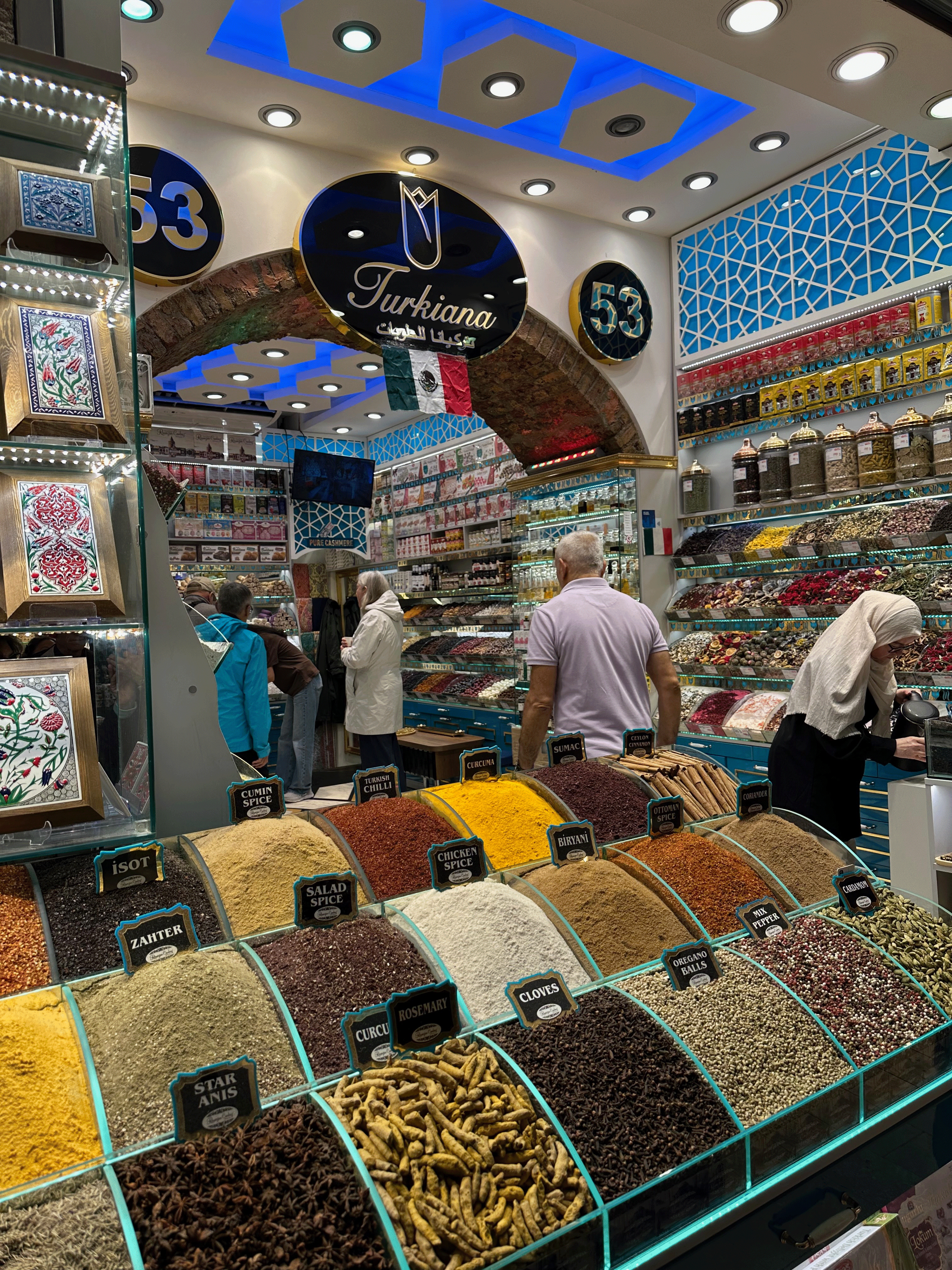 Interior of a spice shop in Istanbul featuring various spices in labeled containers, with customers browsing and colorful displays in the background.