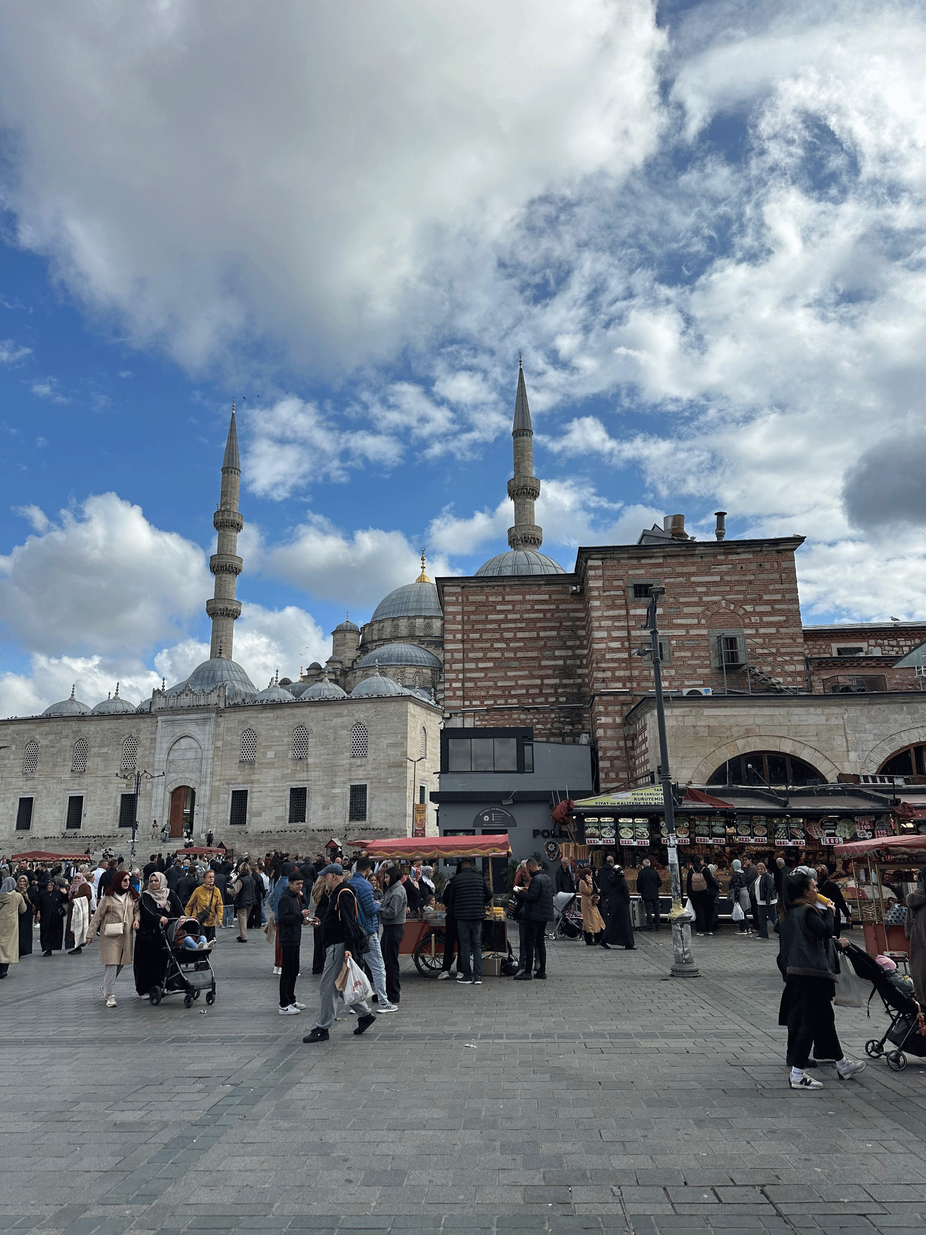 A bustling market square in Istanbul with crowds of people, street vendors, and the domes and minarets of a mosque in the background under a partly cloudy sky.