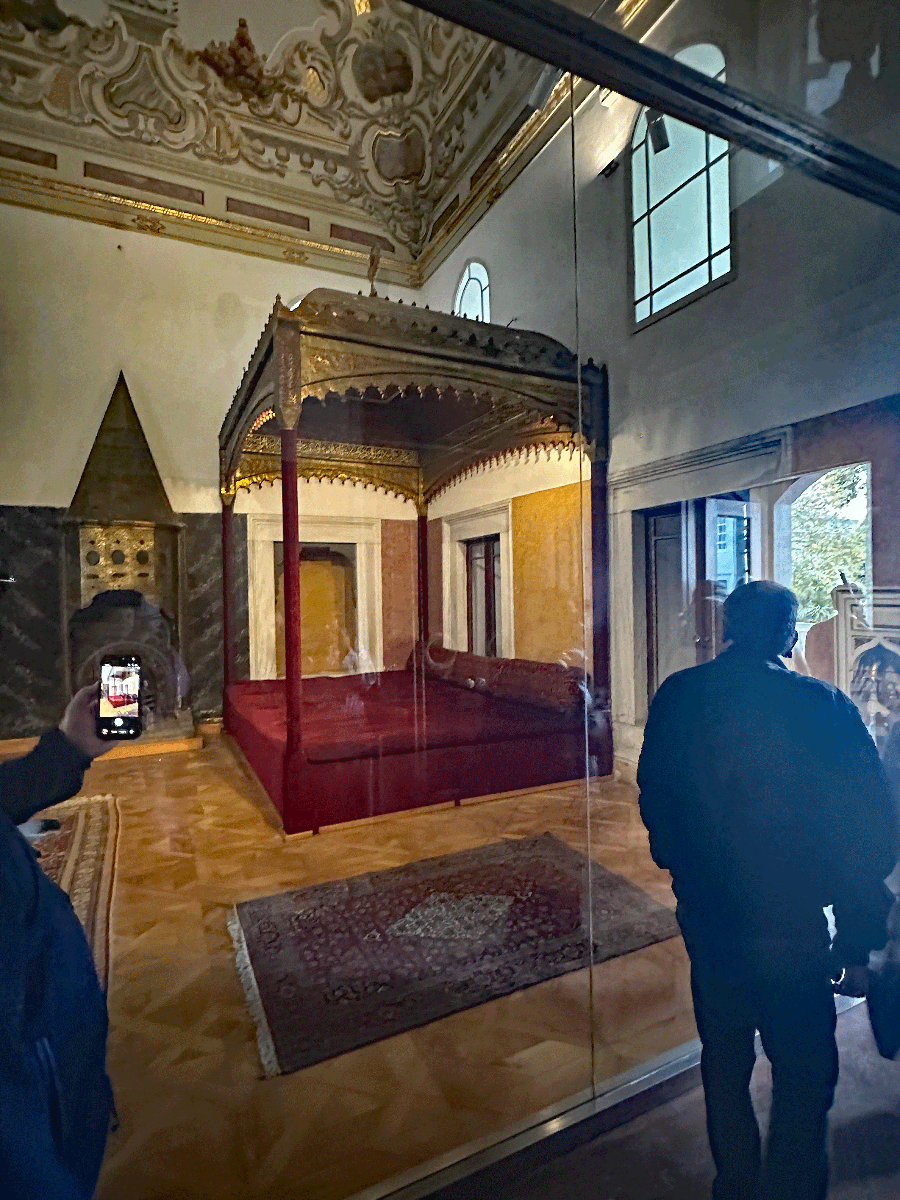 Interior view of a room in Topkapi Palace, featuring an ornate bed canopied with rich fabric and intricate ceiling designs, with visitors reflected in the glass display.