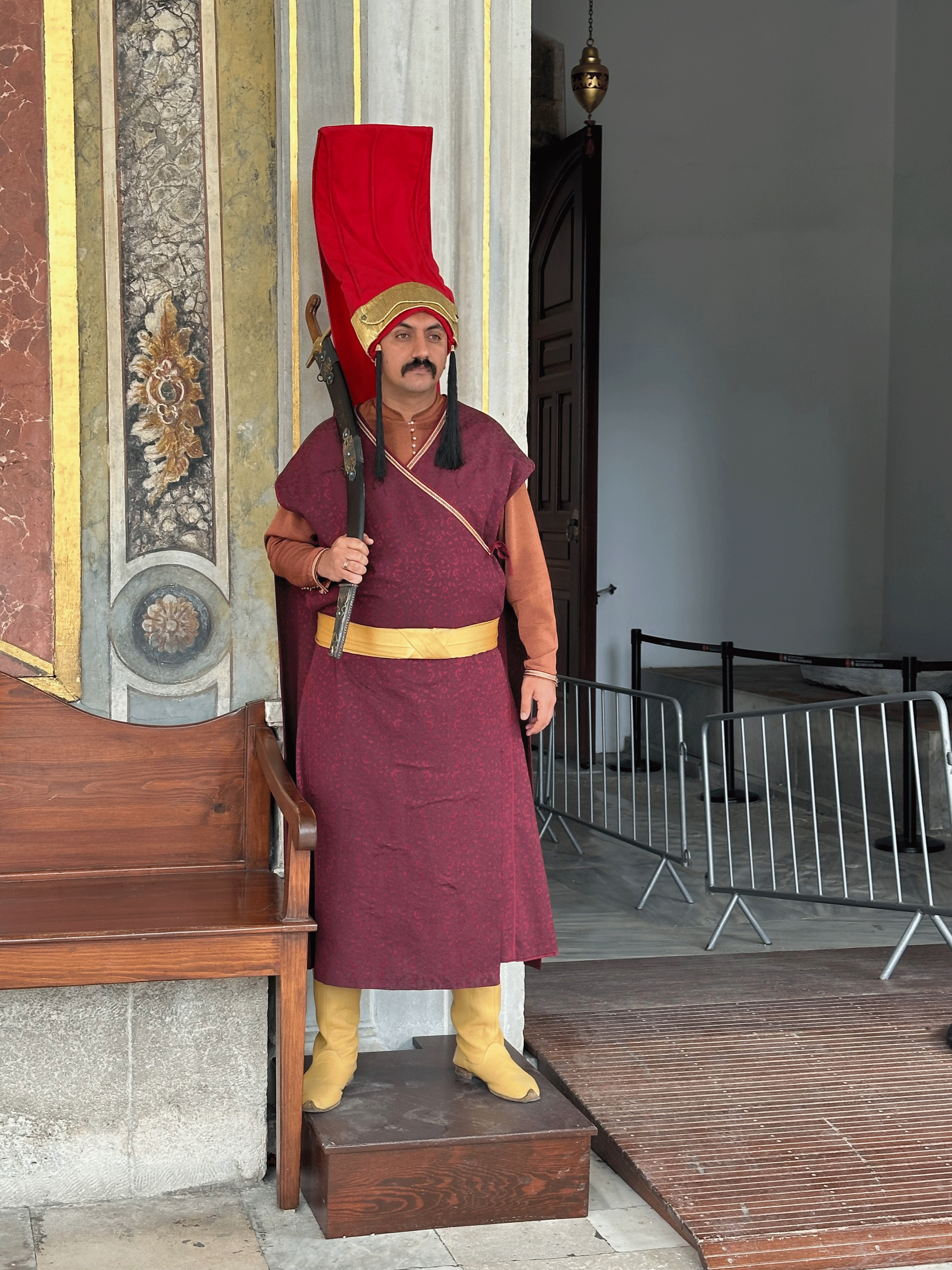 A man dressed in traditional Ottoman guard attire stands at the entrance of a historic building, holding a ceremonial weapon.