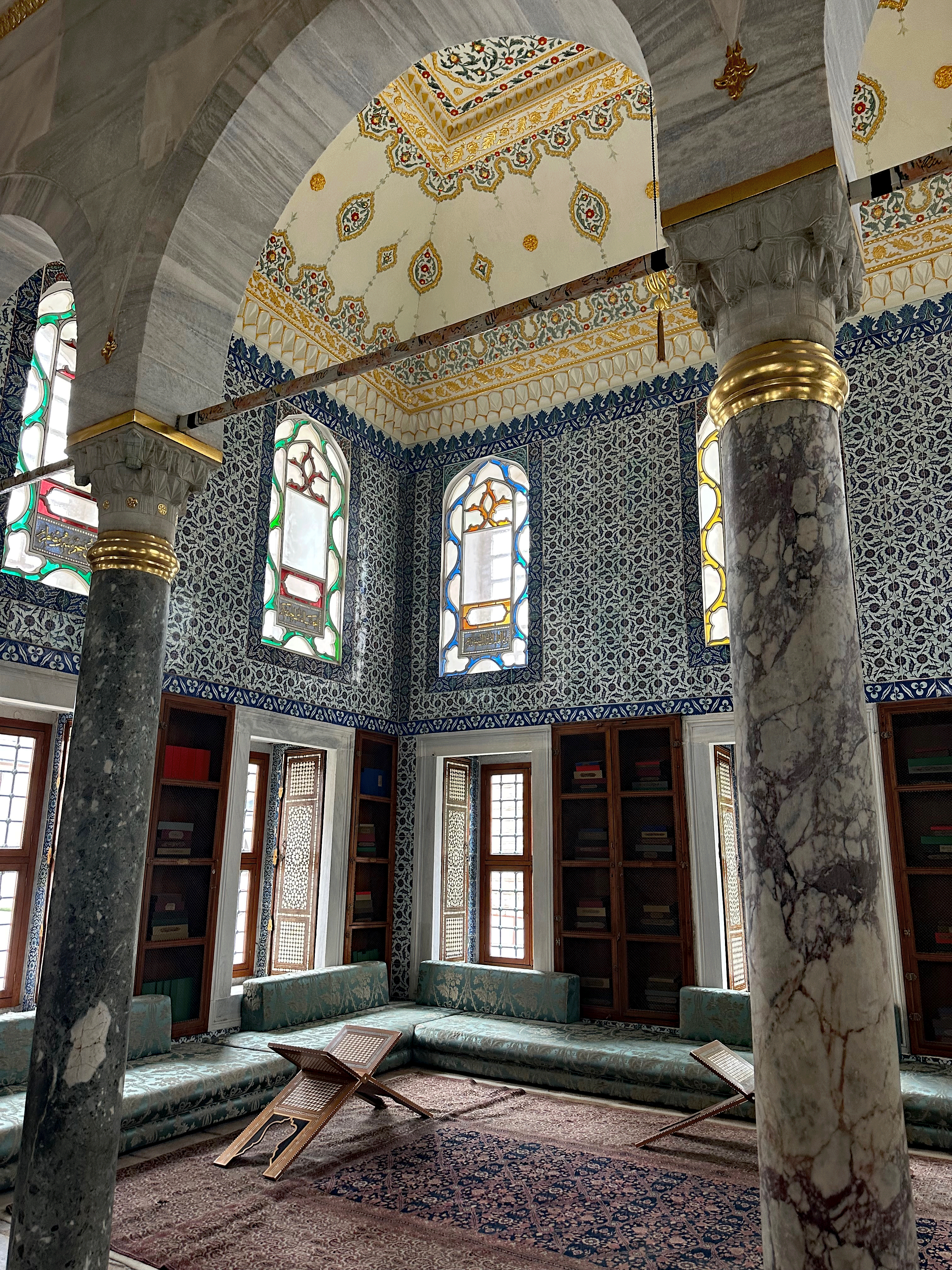 Interior of a historic room in Topkapi Palace, featuring ornate wall patterns, stained glass windows, and traditional Ottoman furniture.
