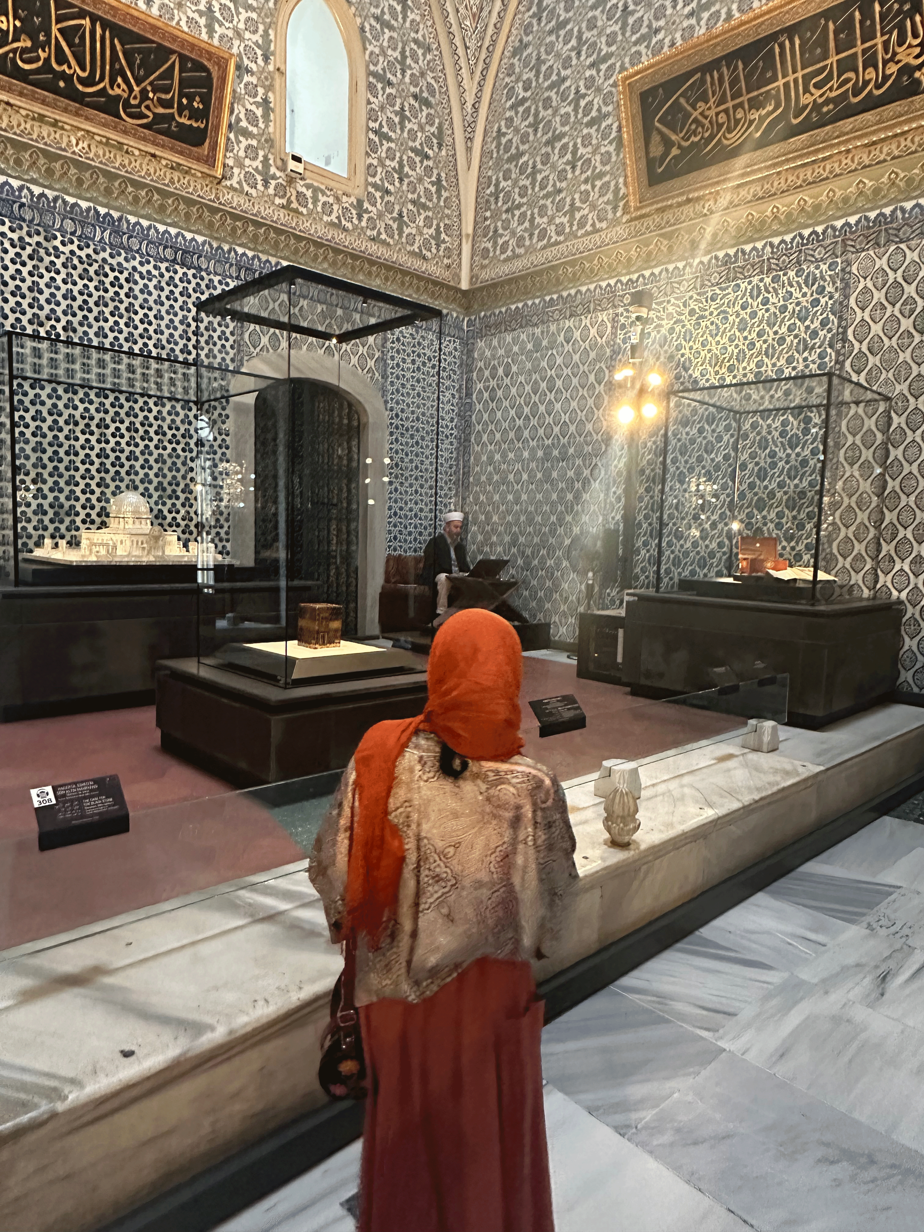 A visitor in traditional dress observing exhibits inside Topkapi Palace, with ornate blue-tiled walls and historical artifacts on display.