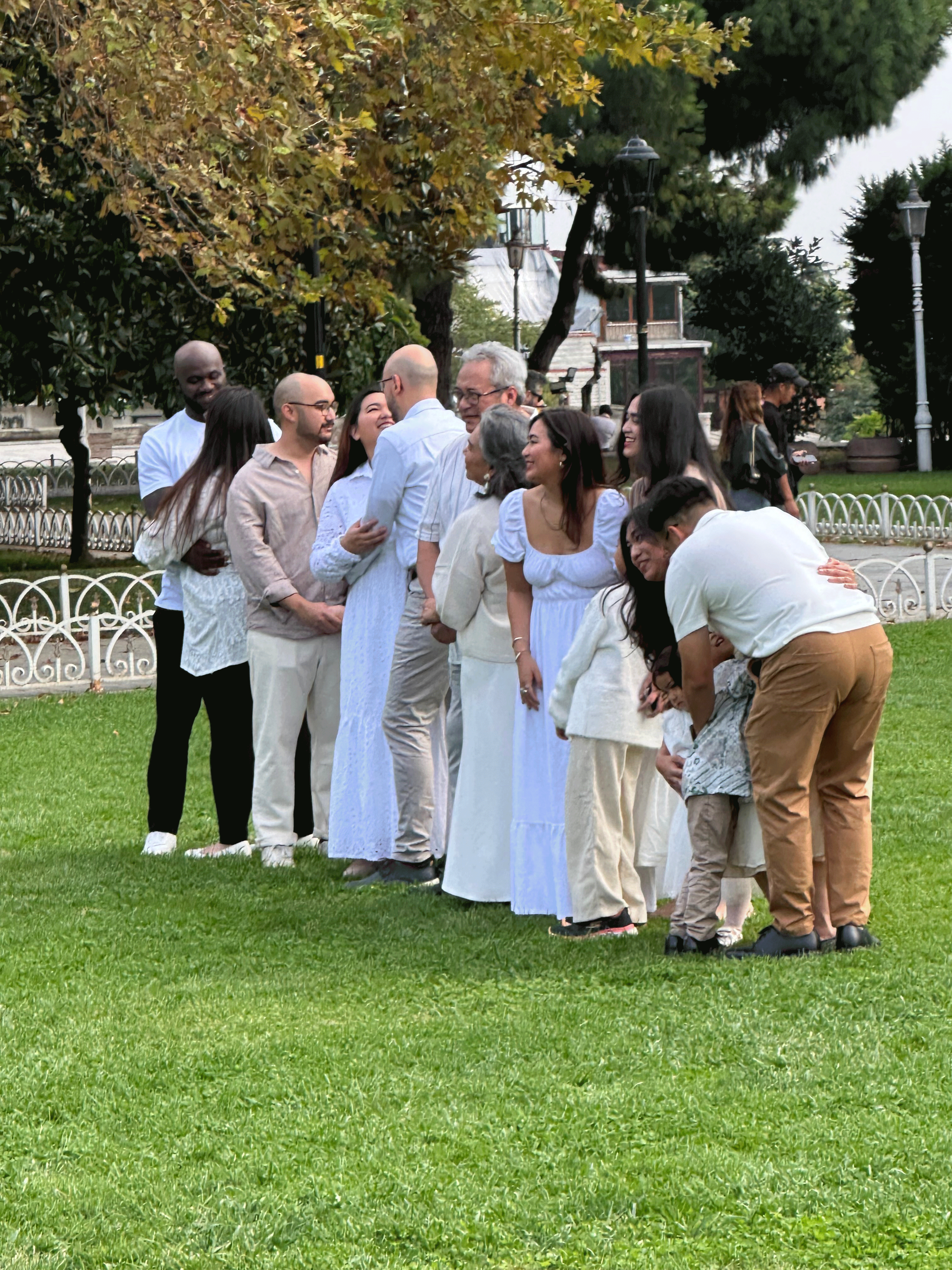 A cheerful group of people in light-colored clothing, posing together on a grassy area with trees in the background, creating a warm and festive atmosphere.