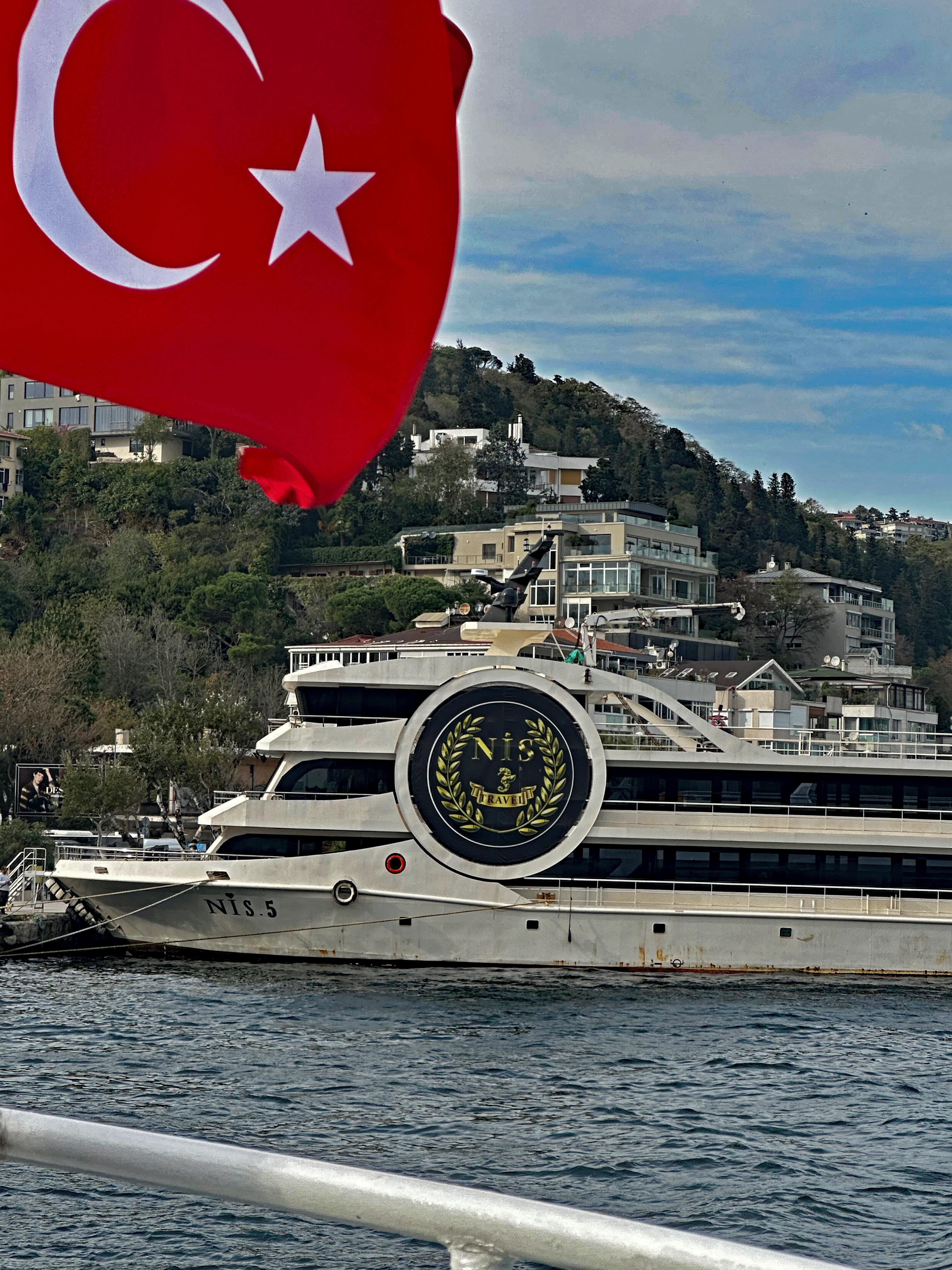 A Turkish flag waving in the foreground with a boat named NIS.5 docked along the Bosphorus Strait, surrounded by lush greenery and modern buildings in Istanbul.