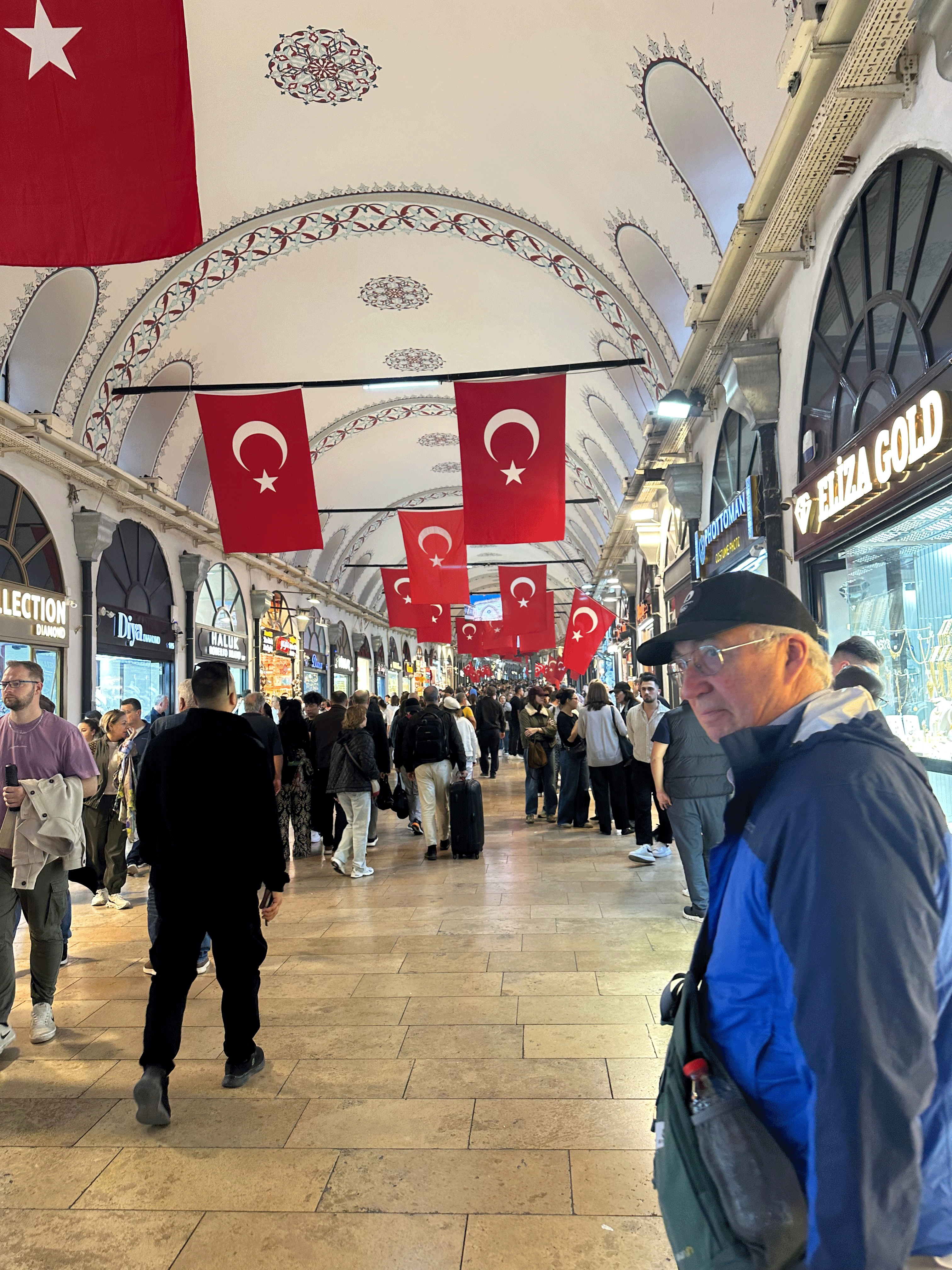 A bustling scene inside the Grand Bazaar in Istanbul, with shoppers exploring shops adorned with Turkish flags hanging from the ceiling.