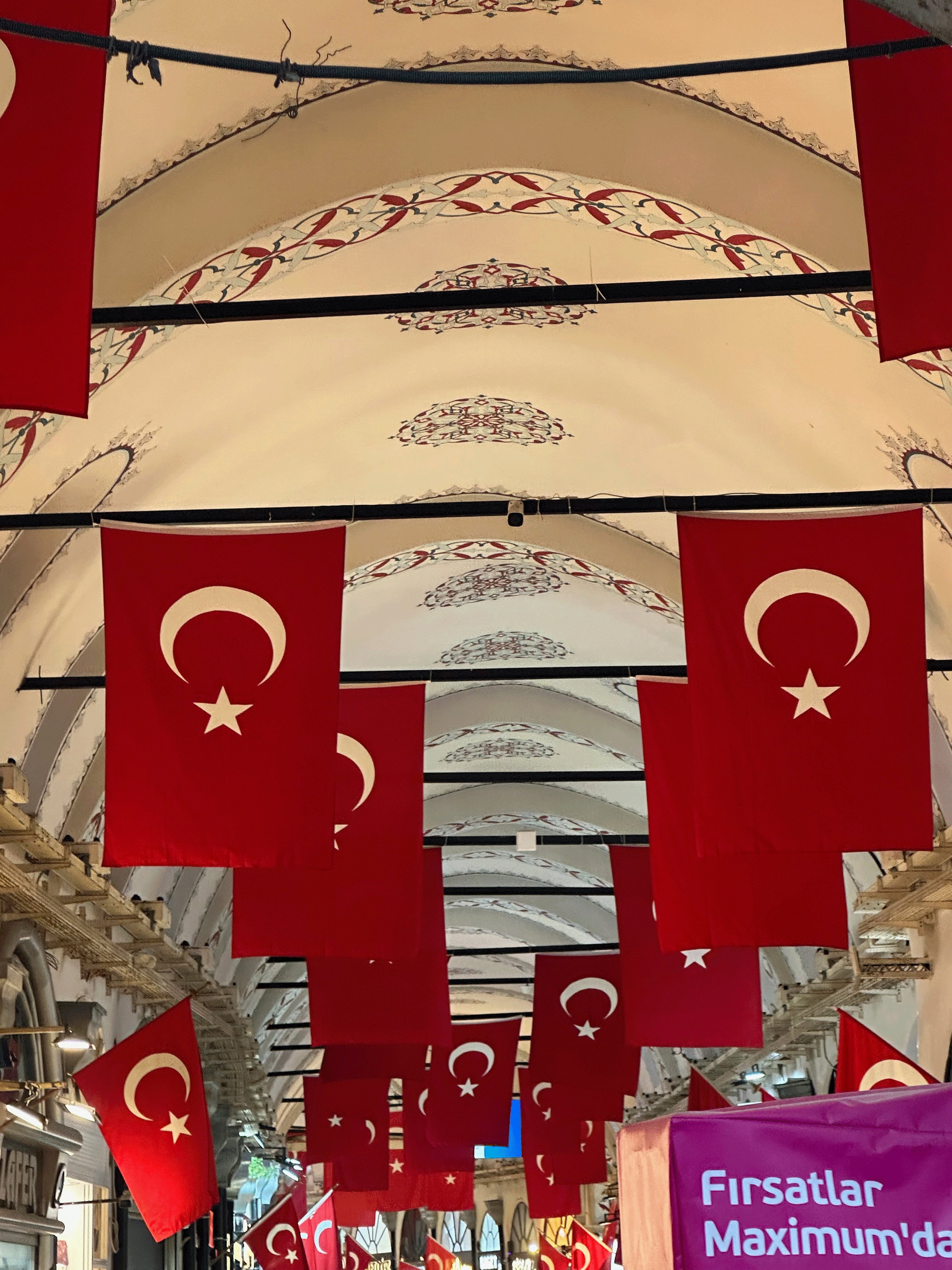 Interior view of a market with Turkish flags hanging from the ceiling, featuring decorative patterns on the roof.