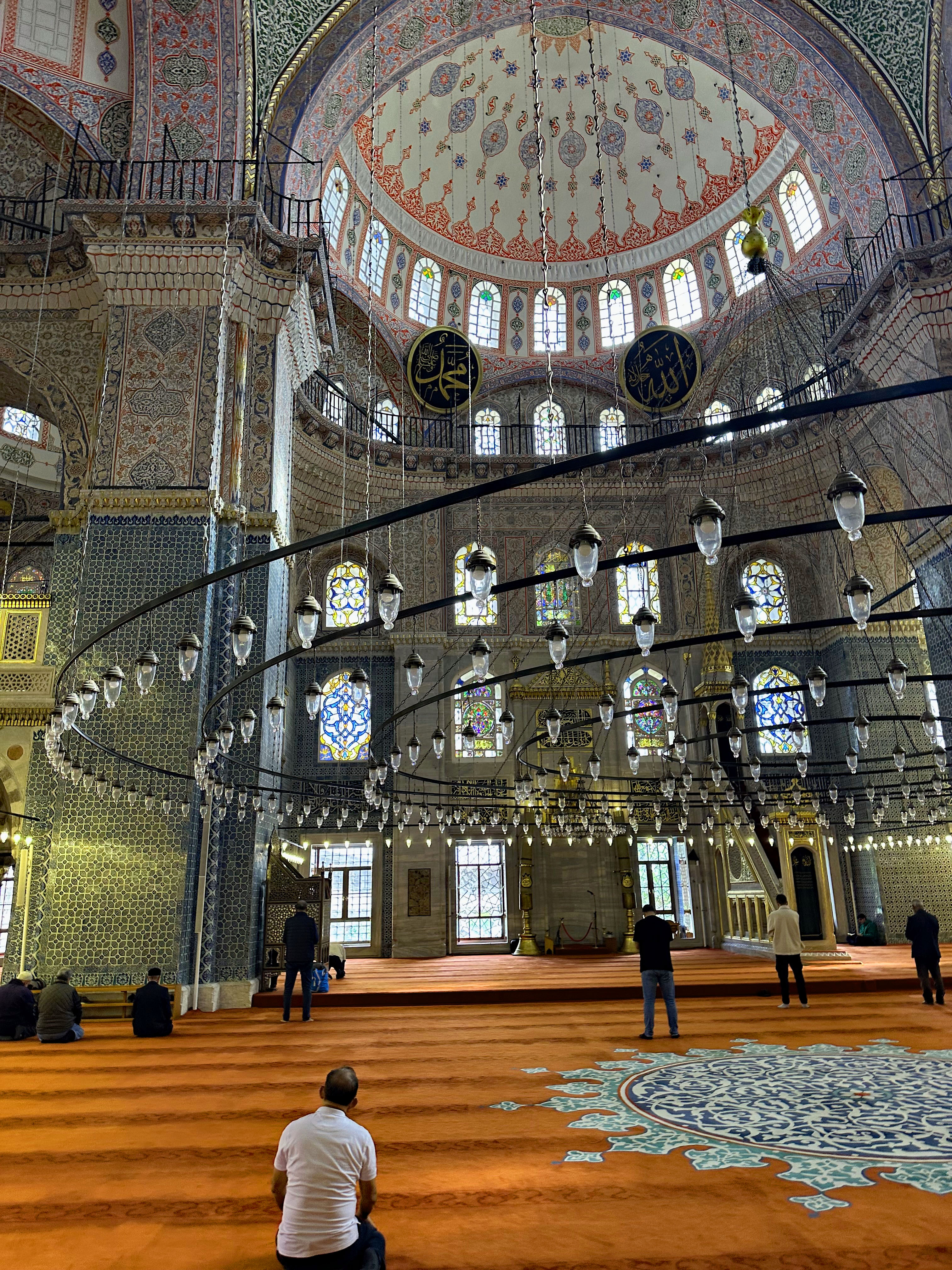 Interior view of the Blue Mosque in Istanbul, featuring ornate decorations, large windows, and worshippers on the orange carpet.