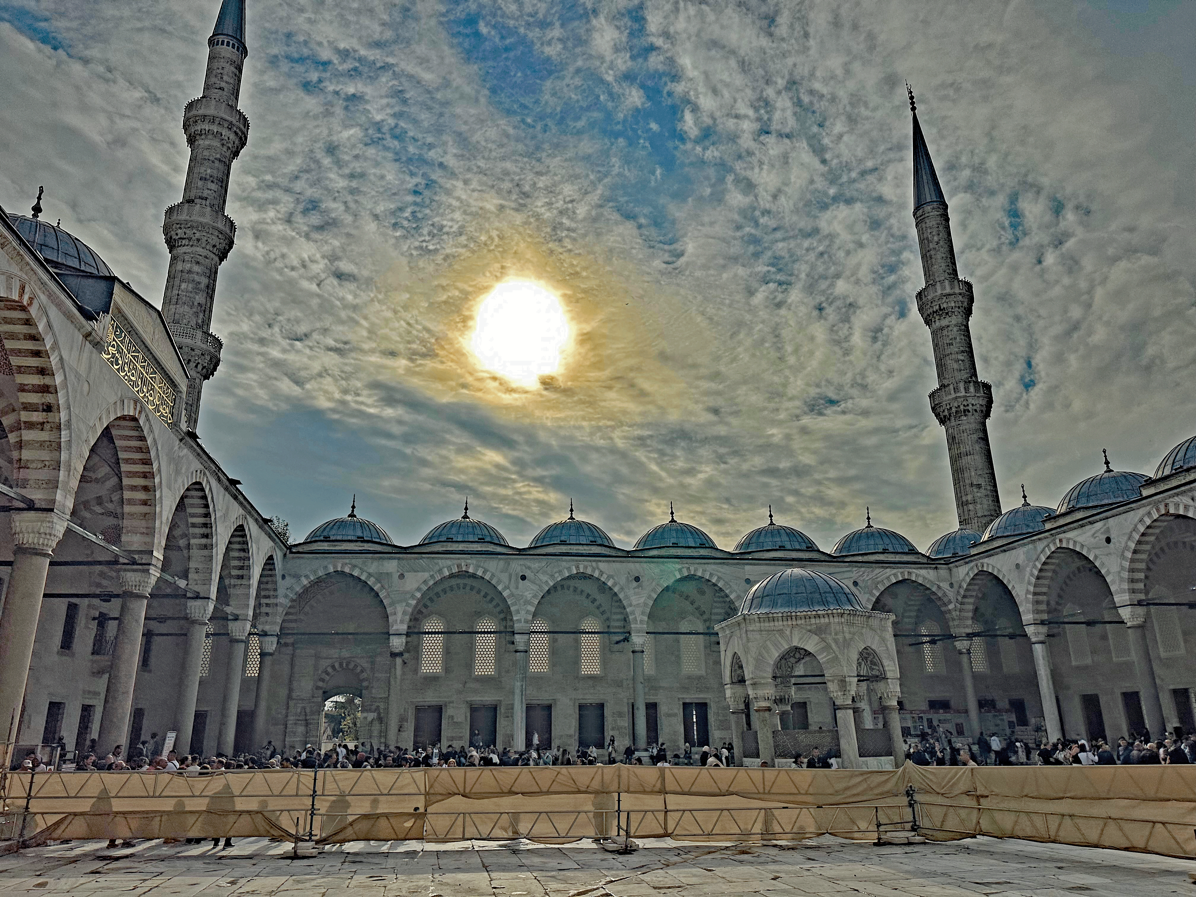 The courtyard of the Blue Mosque in Istanbul, showcasing its magnificent architecture with six minarets and domed roofs under a cloudy sky.