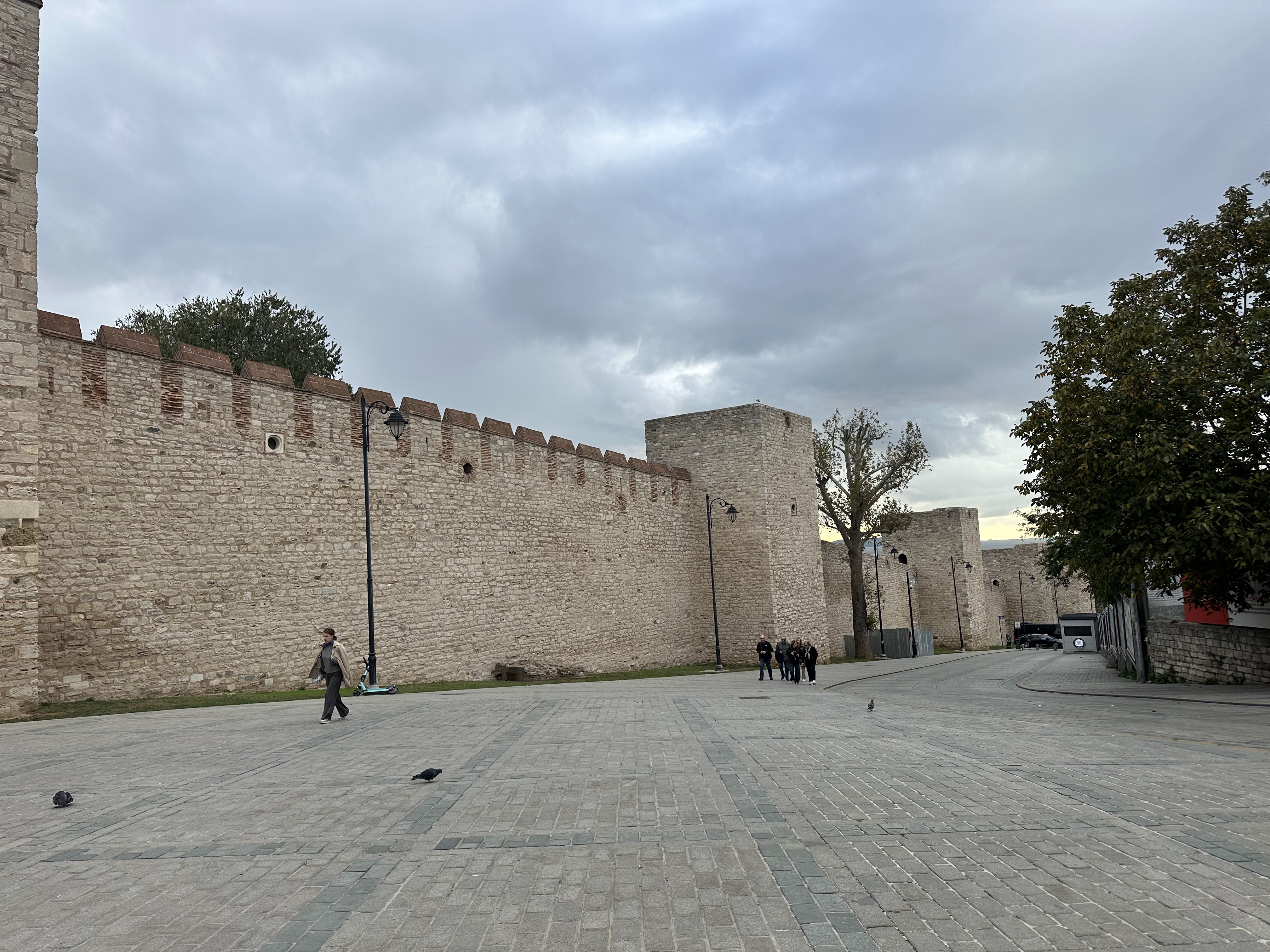 A stone wall with towers along an empty street in Istanbul, with a cloudy sky above and a few people walking nearby.