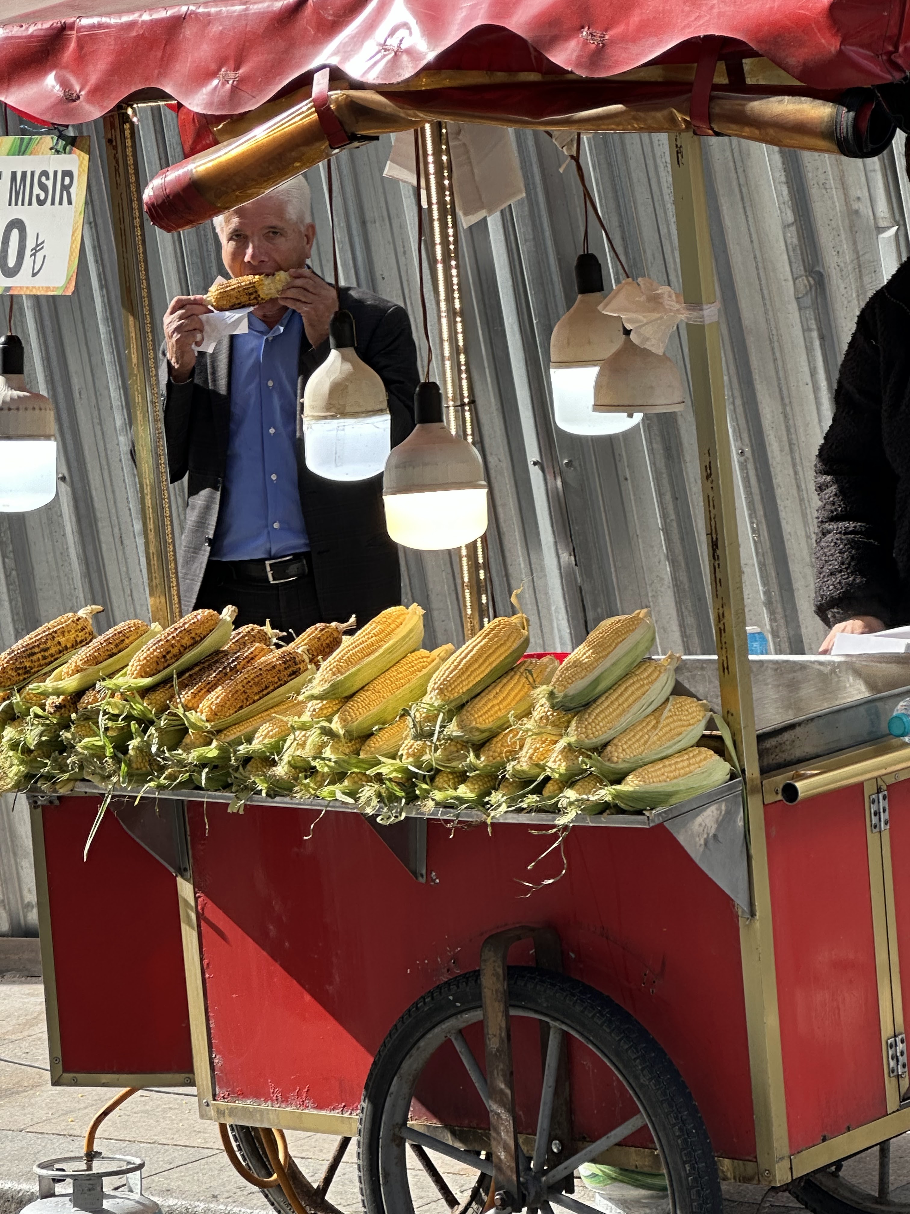 A street vendor selling grilled corn on a cart in Istanbul, with a man enjoying an ear of corn in front of the vendor's stall, which has a red exterior and hanging lights.