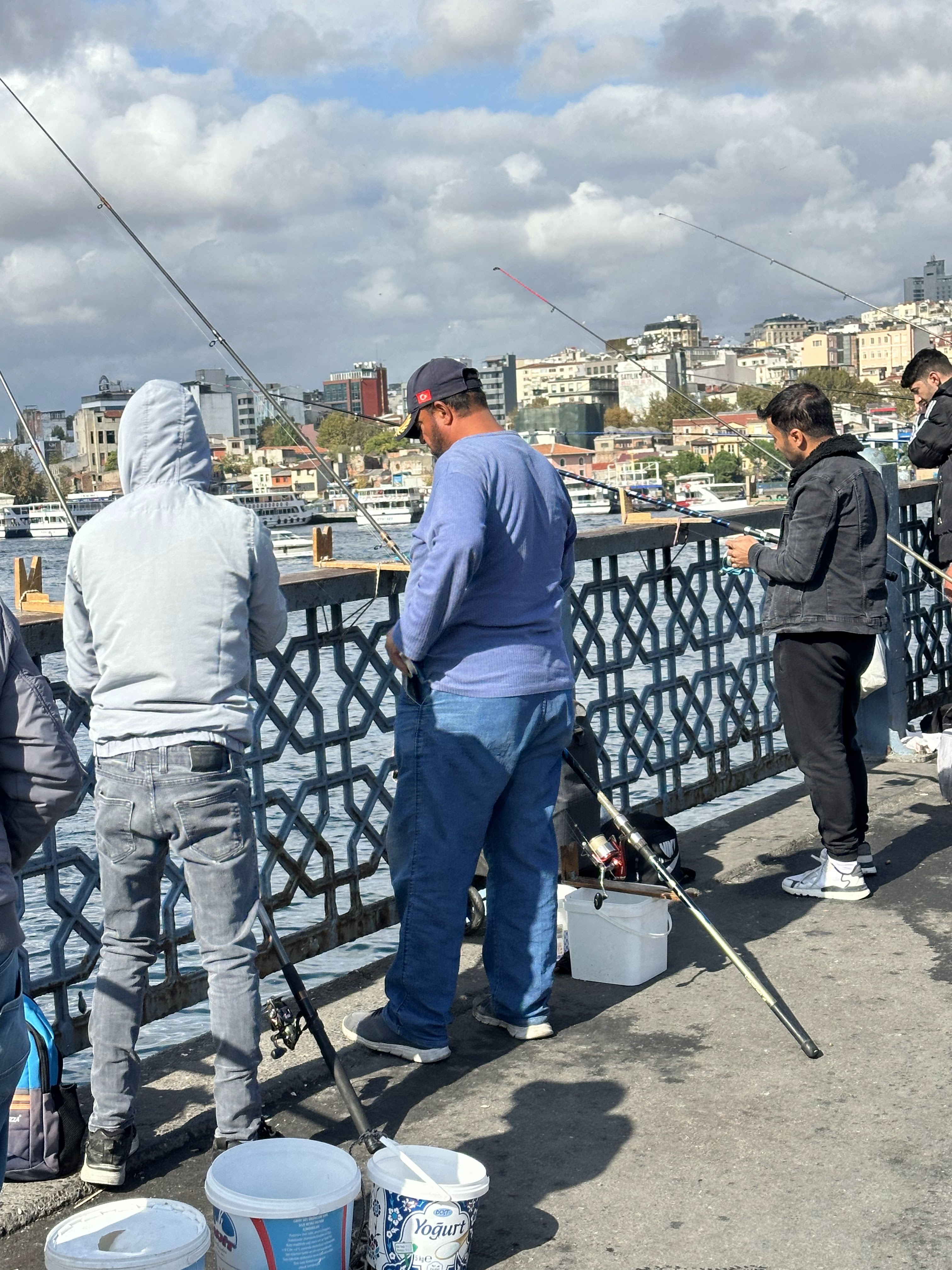 A group of people fishing from a bridge in Istanbul, with fishing rods and buckets nearby, against a backdrop of urban buildings and a cloudy sky.